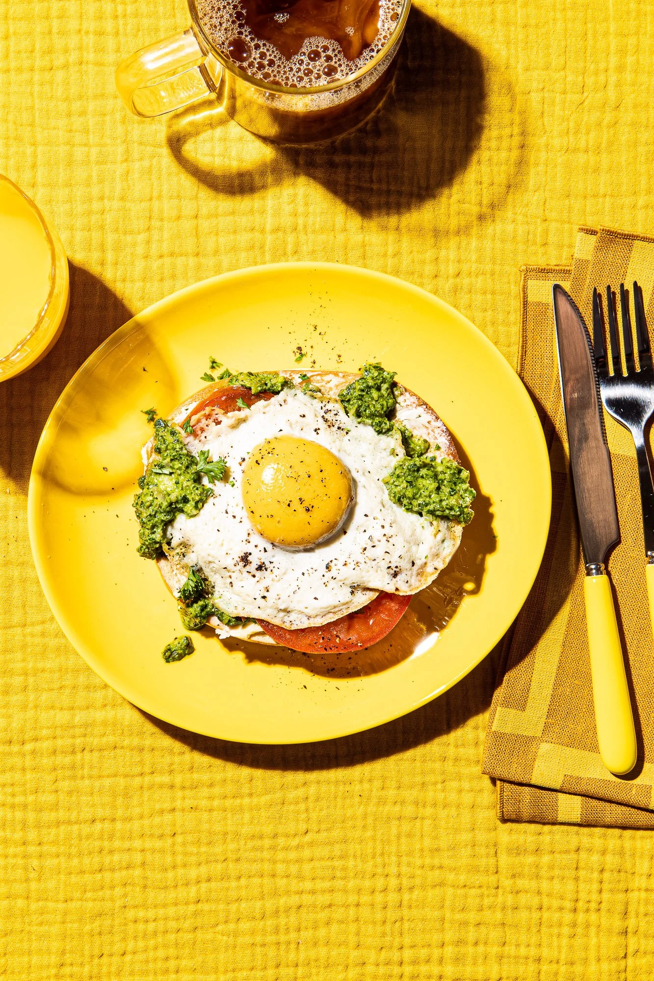 A yellow plate with a breakfast dish of a vegan fried egg, tomato slices, and green pesto on top, placed on a yellow tablecloth. To the side is a fork and knife on a yellow napkin, with a glass cup of coffee.