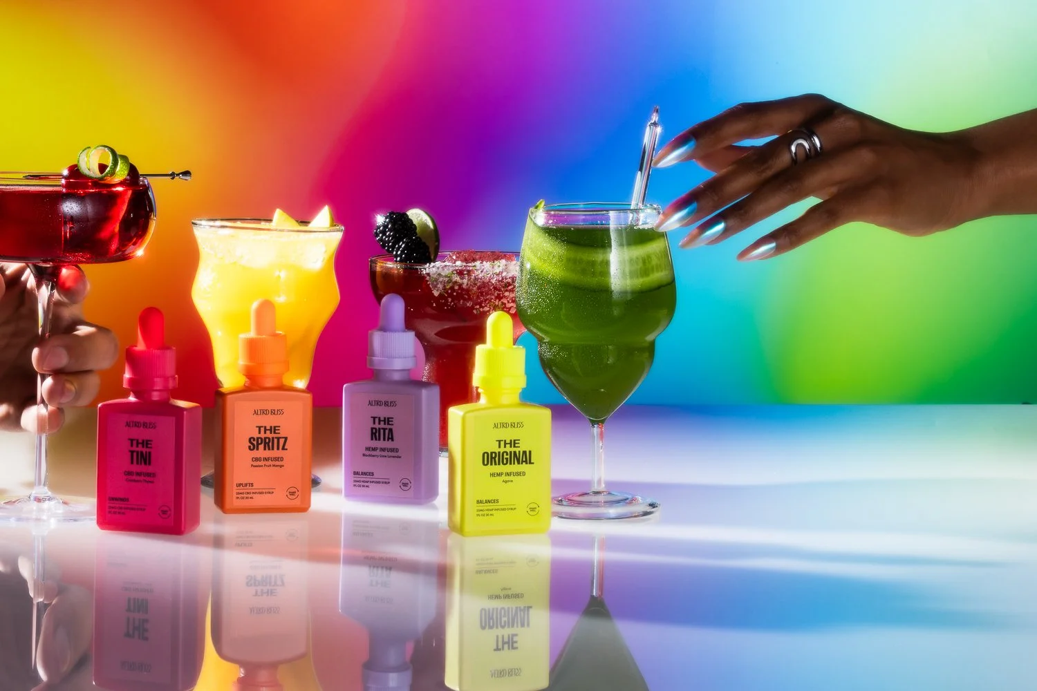 Colorful cocktails and CBD oil bottles on a reflective surface with a rainbow background.