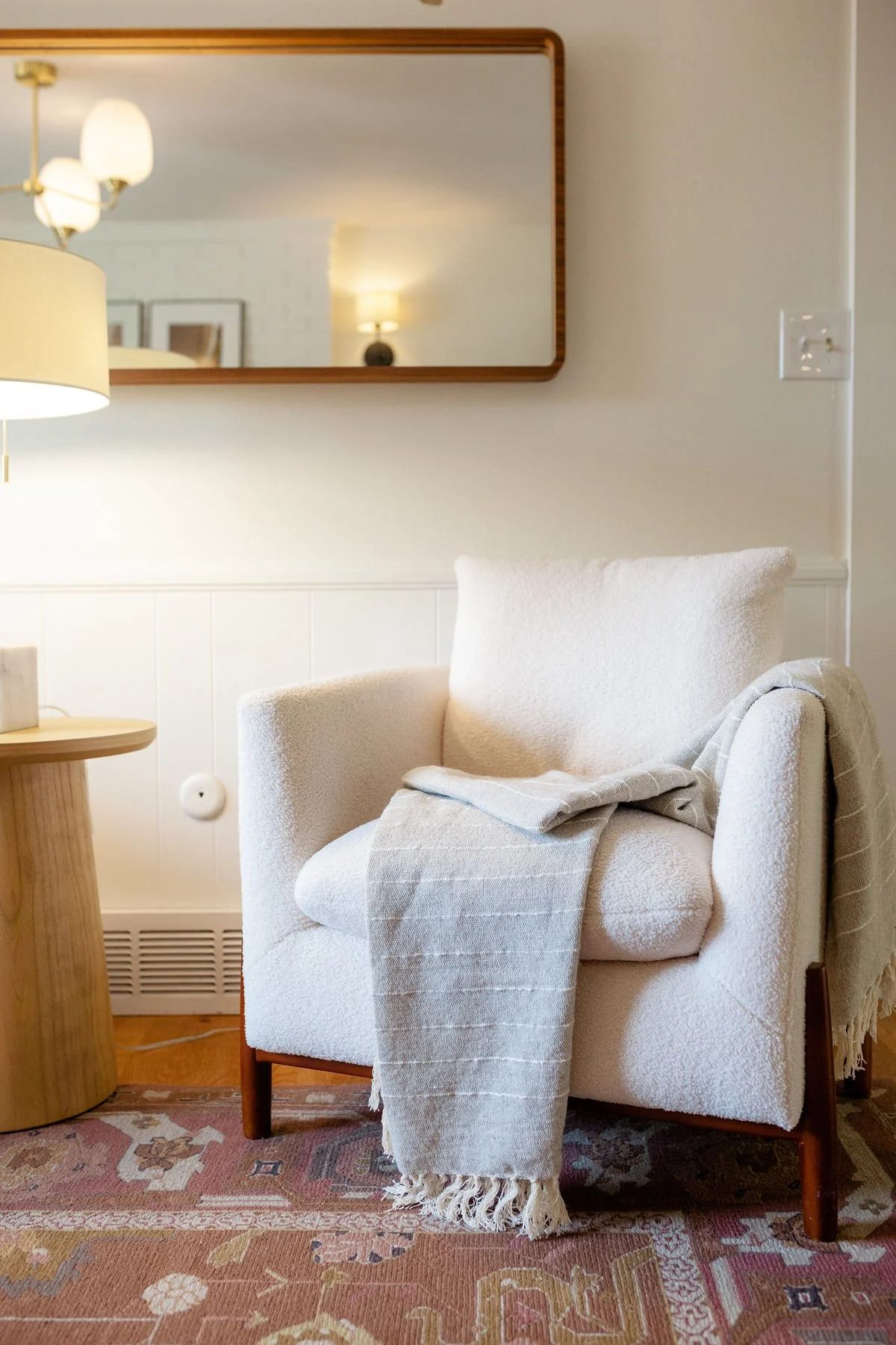 A cozy living room corner with a cream upholstered armchair draped with a beige striped throw blanket, a round wooden side table with a lamp, and a large mirror above reflecting a wall with framed pictures and additional lighting.