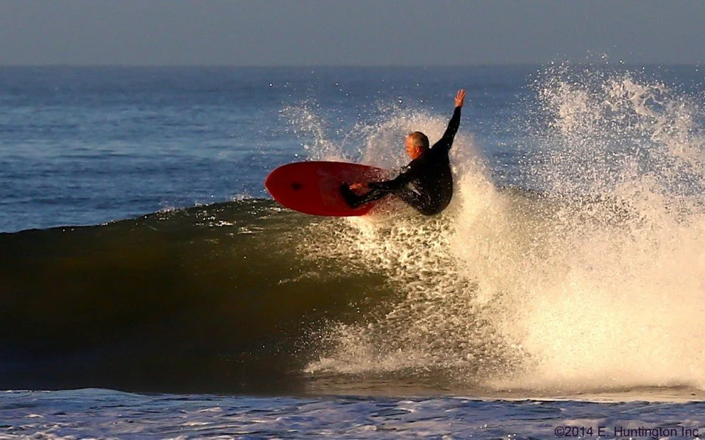 A person in a black wetsuit surfing on a red surfboard on a wave at the beach.