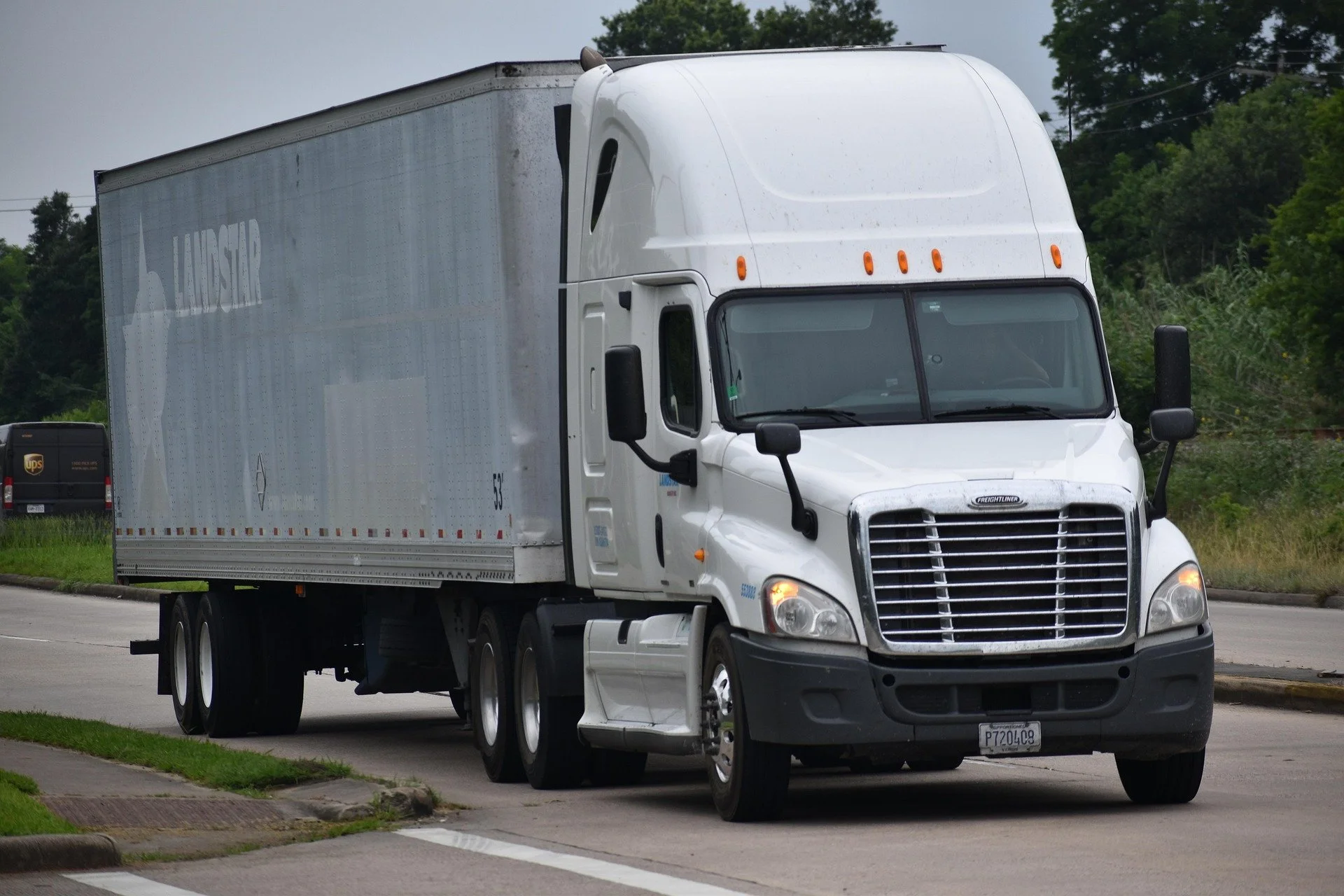 White semi-truck driving on a road, with a gray trailer and trees in the background.