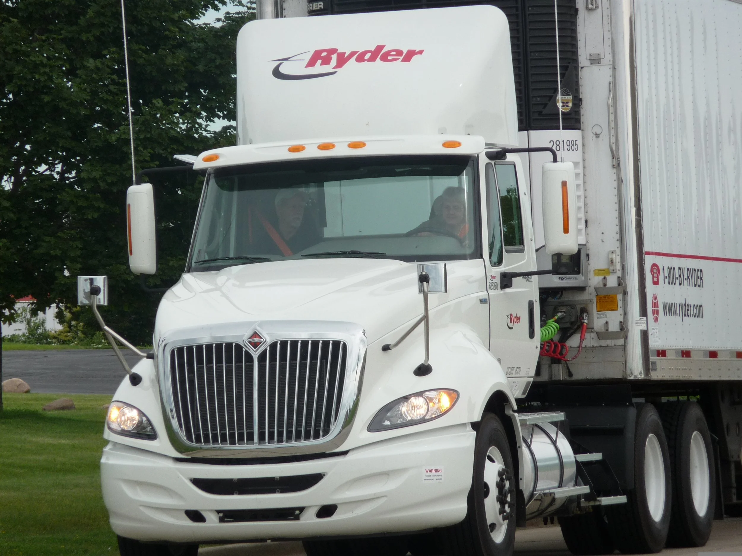 White Ryder semi-truck with a large front grille, headlights on, and a cab with two drivers inside, on a paved area with trees in the background.