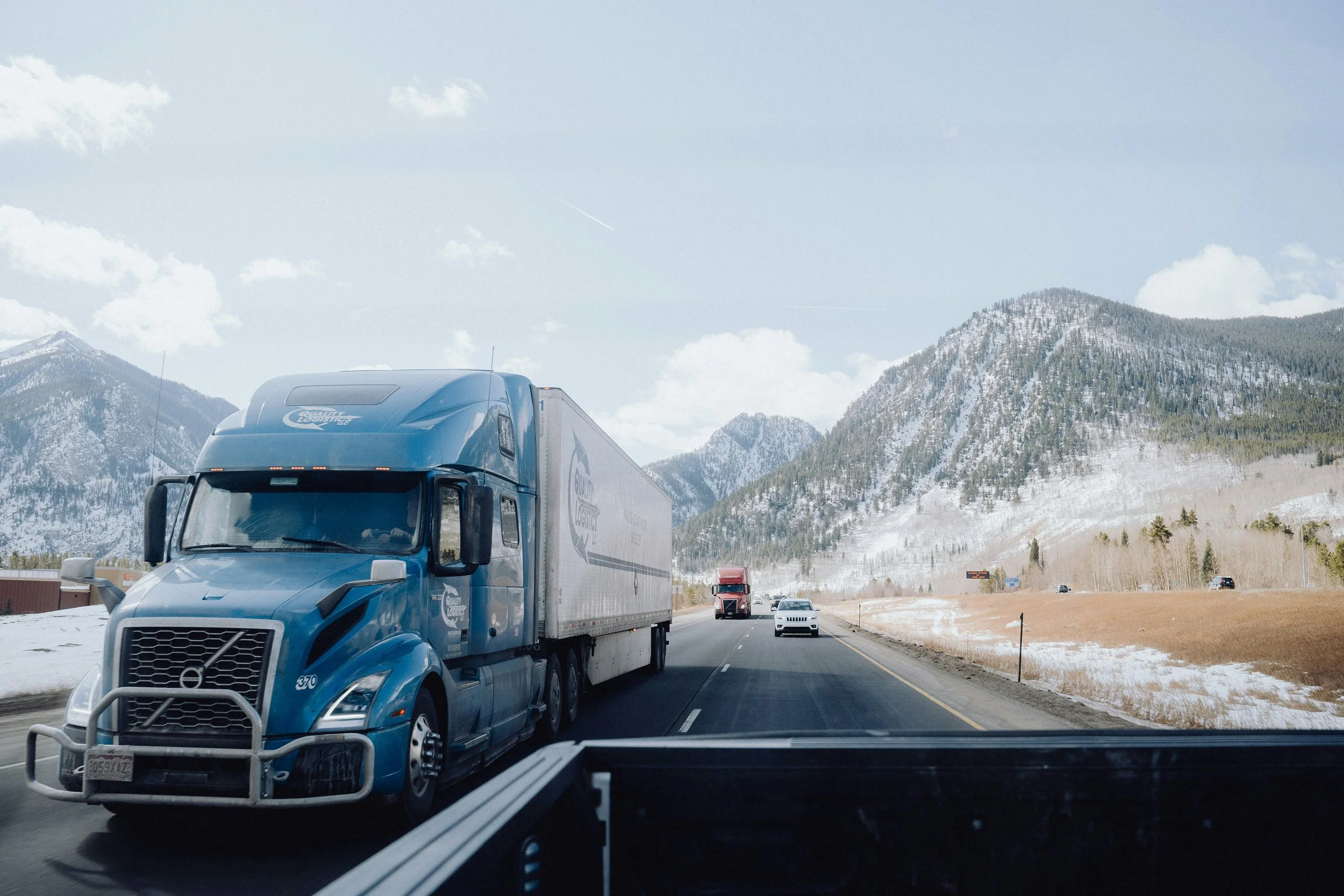 A blue semi-truck driving on a highway with snow-covered mountains and blue sky in the background.