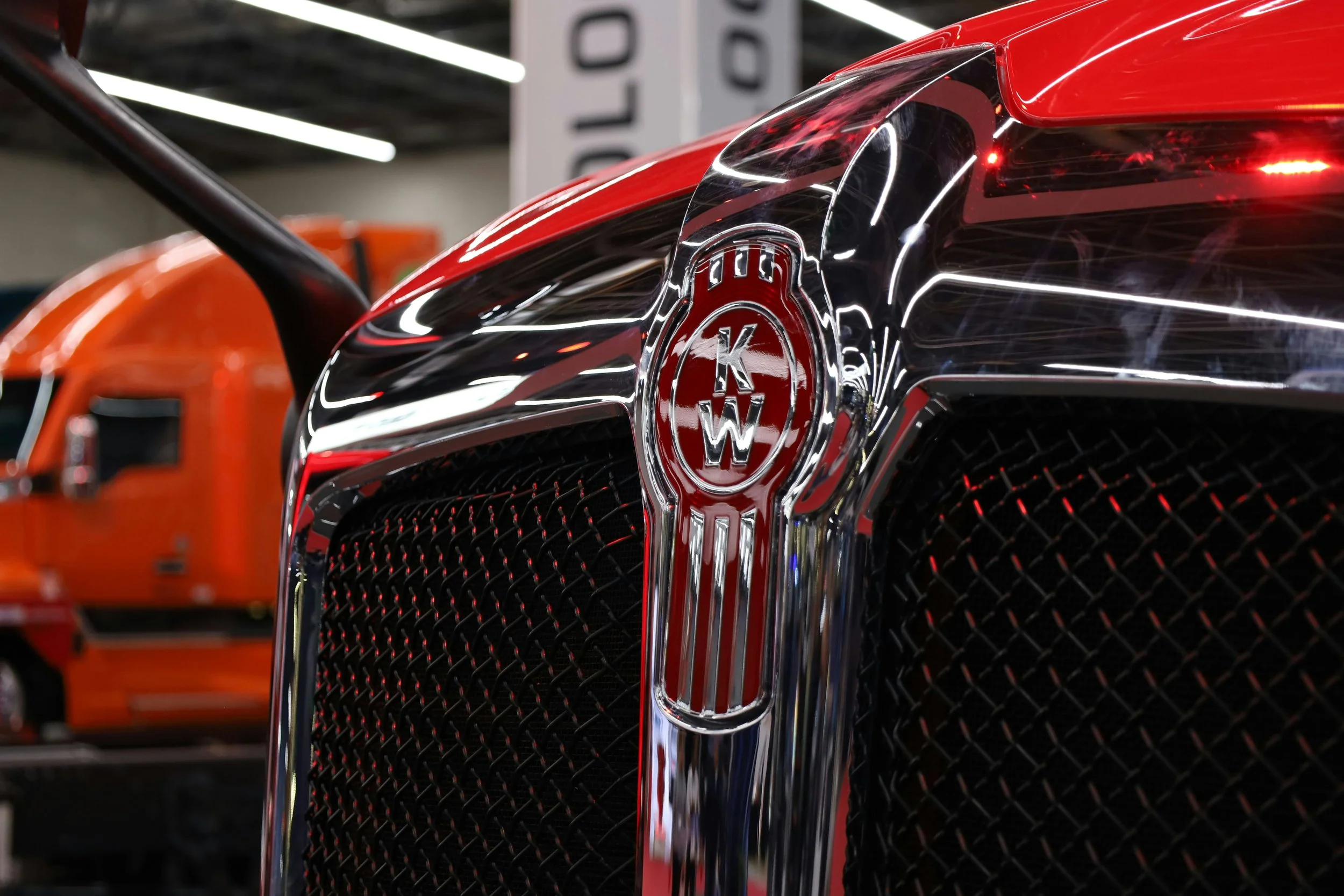 Close-up of the front grille of a red Kenworth semi-truck, with an orange semi-truck in the background inside a showroom or storage facility.