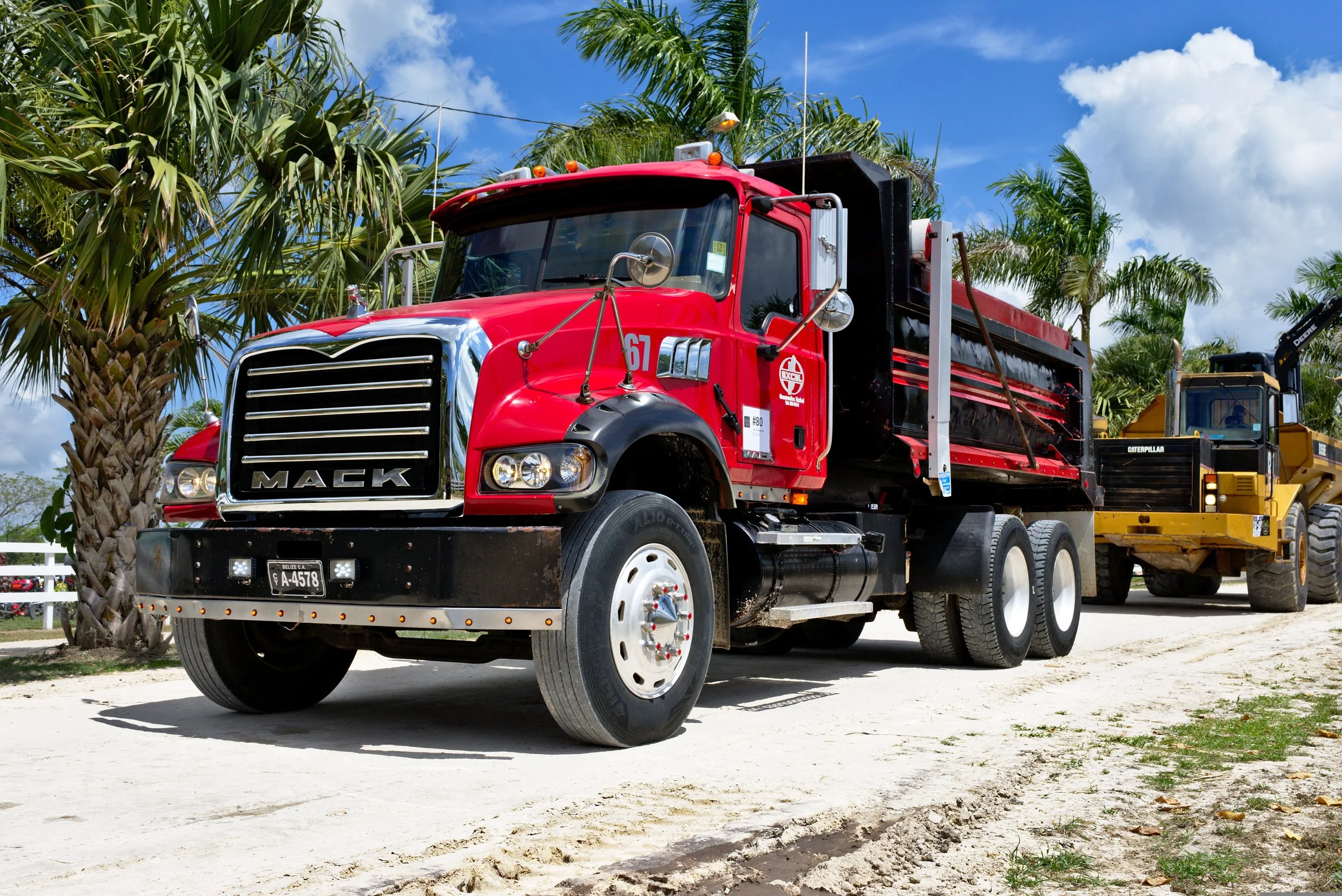 A red Mack fire truck parked on a dirt road, with green palm trees and blue sky in the background.