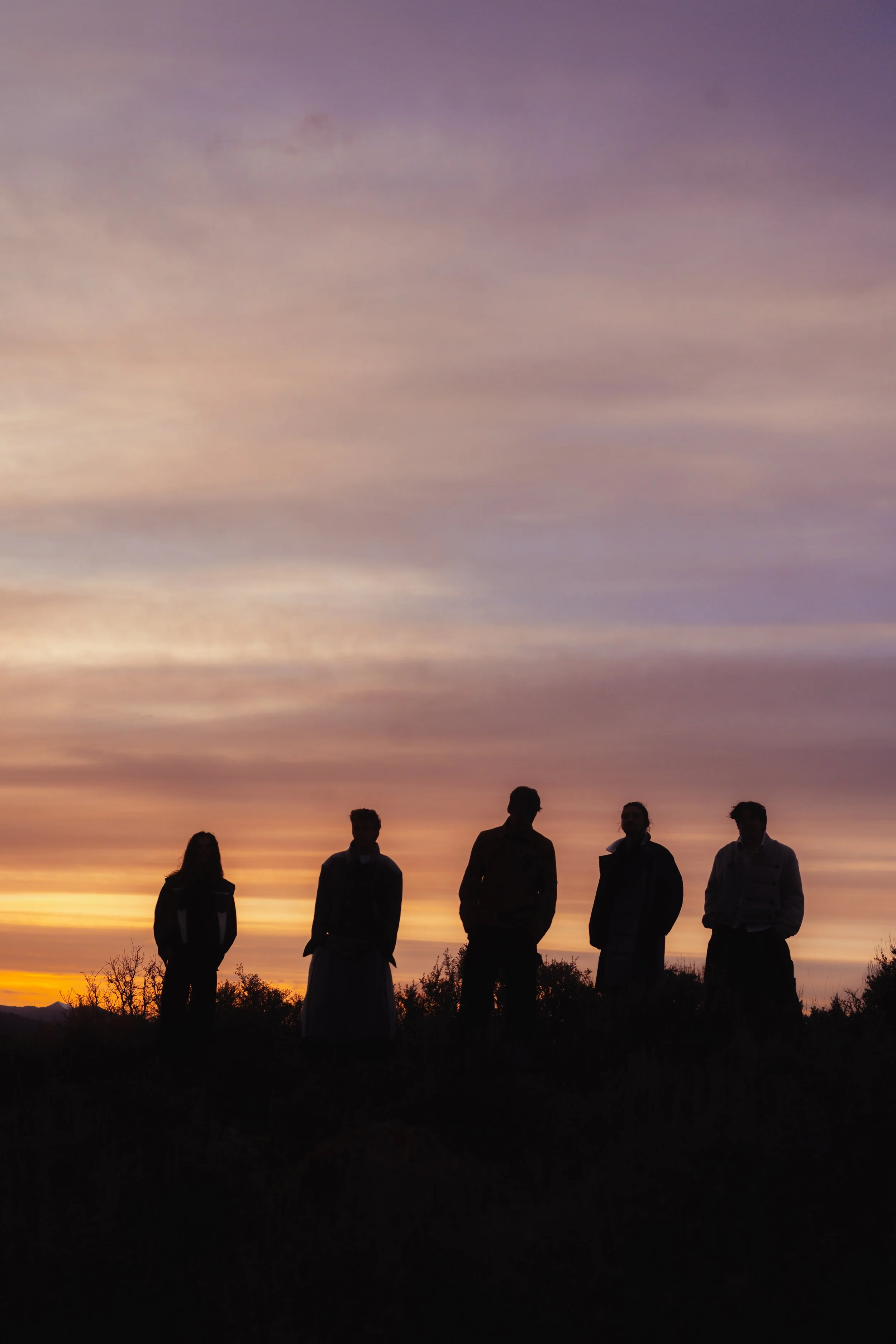 Silhouettes of five people standing outdoors during sunset with a colorful cloudy sky and trees in the background.