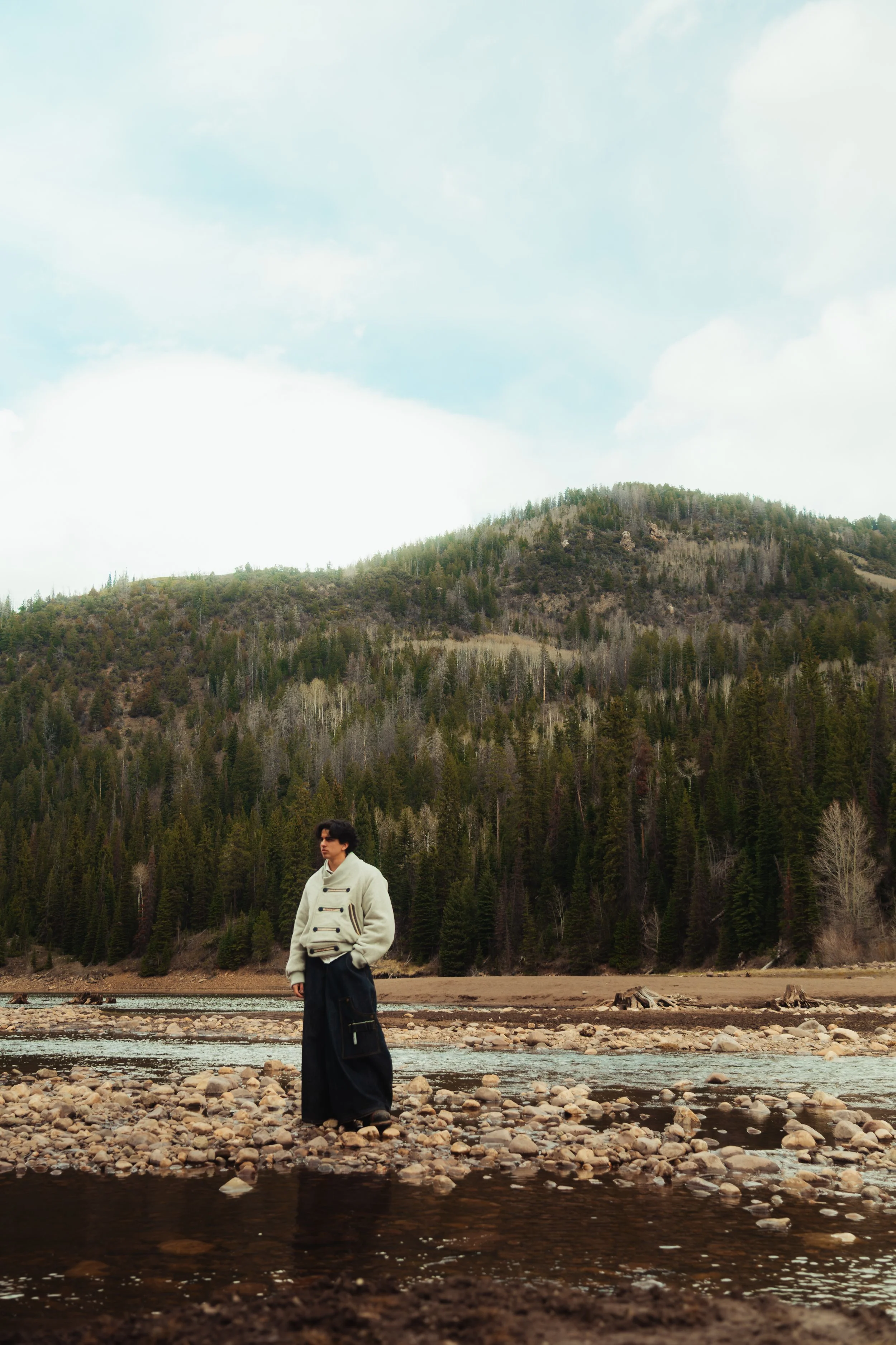 A person standing in a shallow river with a mountainous forested landscape in the background.