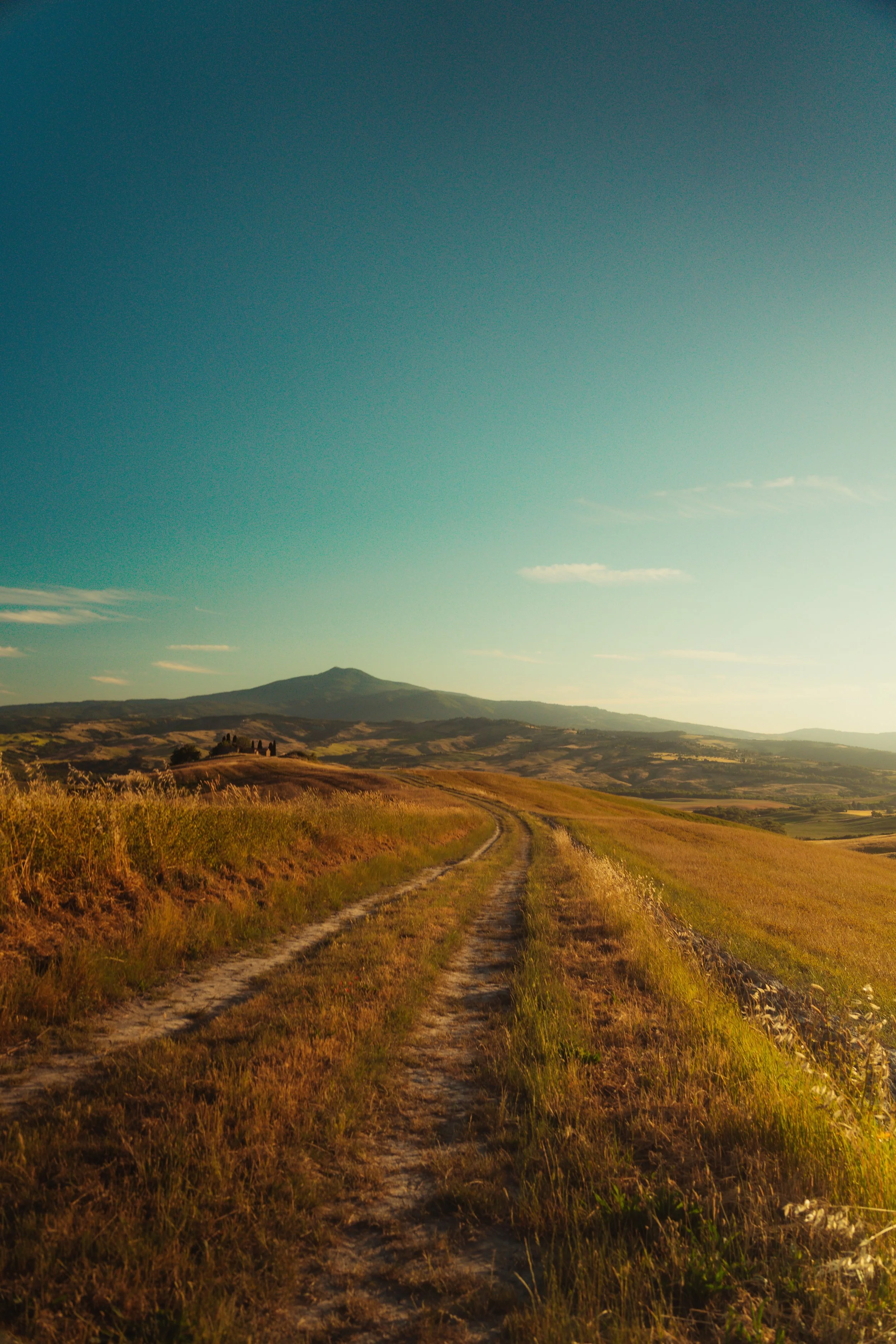 A dirt road runs through grassy fields with rolling hills and mountains in the distance under a clear, teal sky.