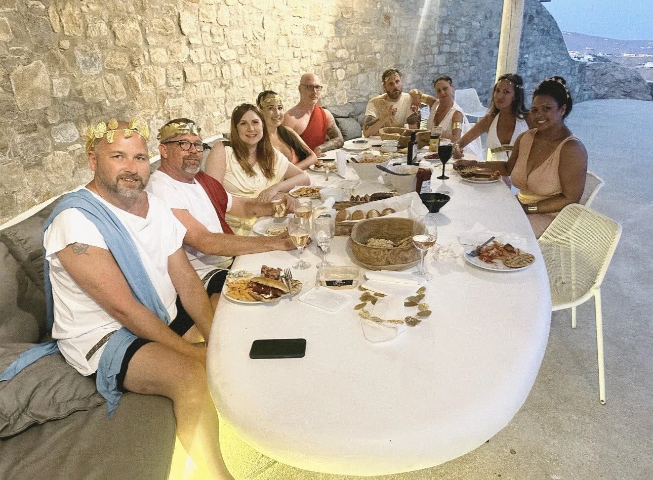 A group of people wearing togas sit around a white table in front of a stone wall.
