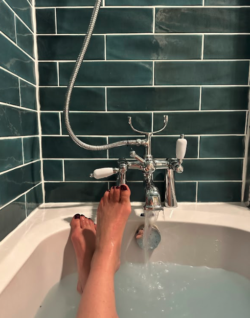 A woman's feet on the edge of a running bathtub, against dark teal-green tile.