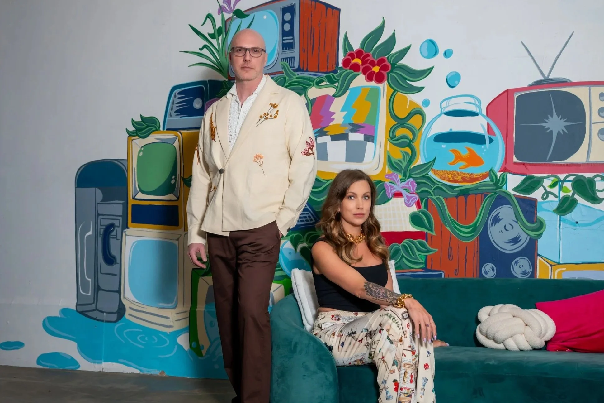 A man standing and a woman sitting in front of a colorful mural featuring old televisions, plants, and fish bowls.