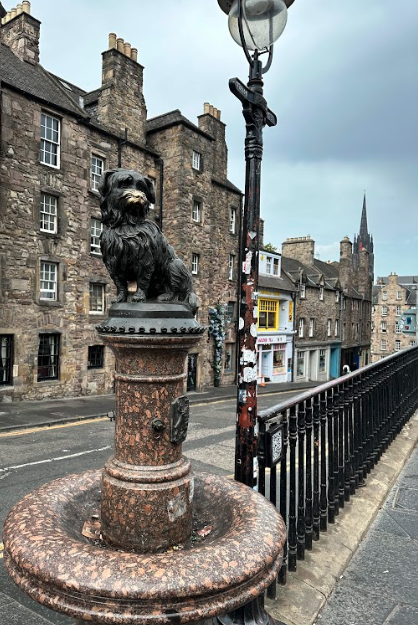 Granite fountain with a bronze statue of a dog on top of it, in front of stone buildings.