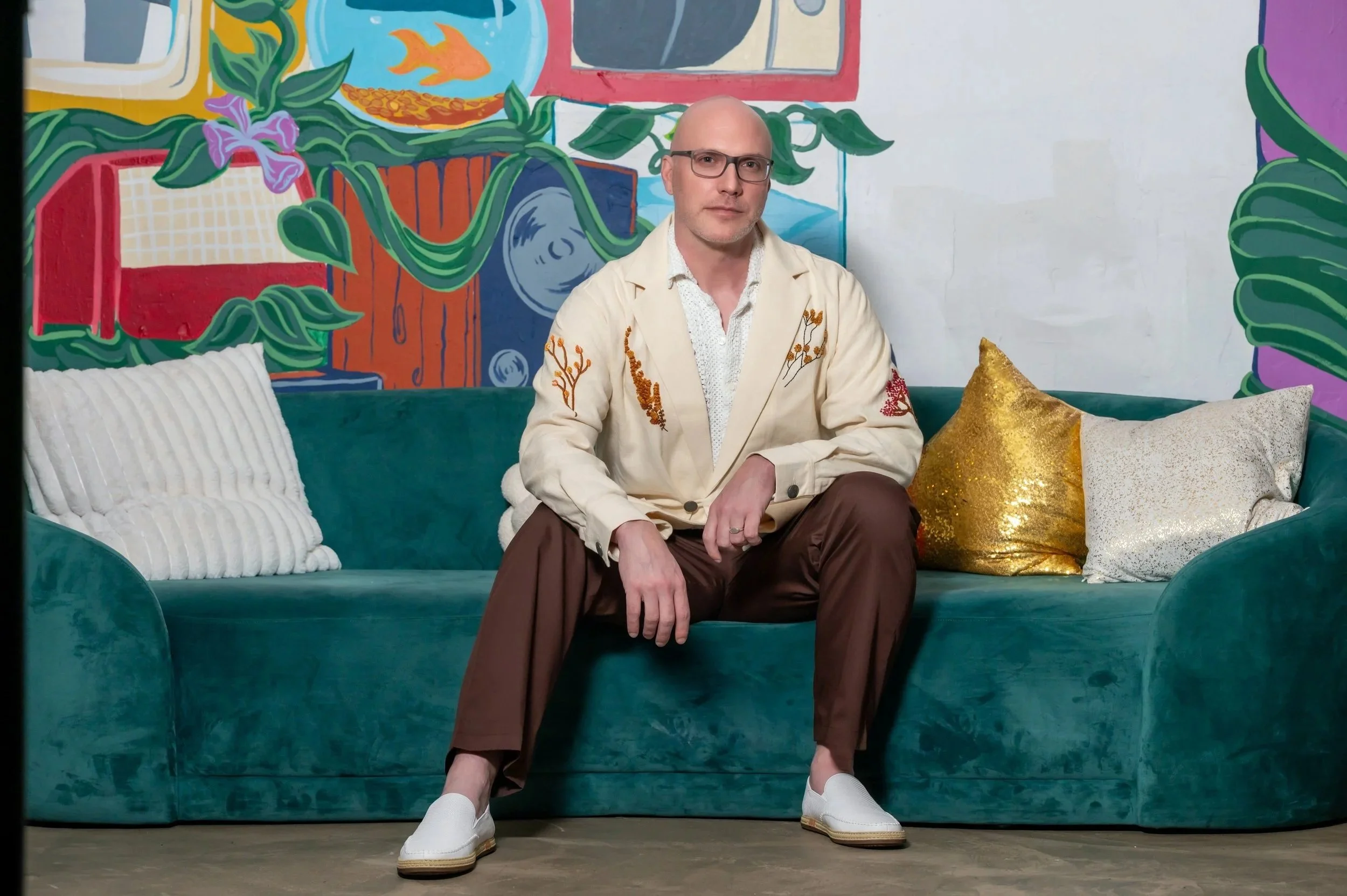 A man with glasses, bald head, dressed in a cream blazer with floral embroidery, white shirt, brown pants, and white slip-on shoes, sitting on a teal velvet sofa with three cushions—white, gold, and silver—against a colorful, abstract mural background with a potted plant and fish design.