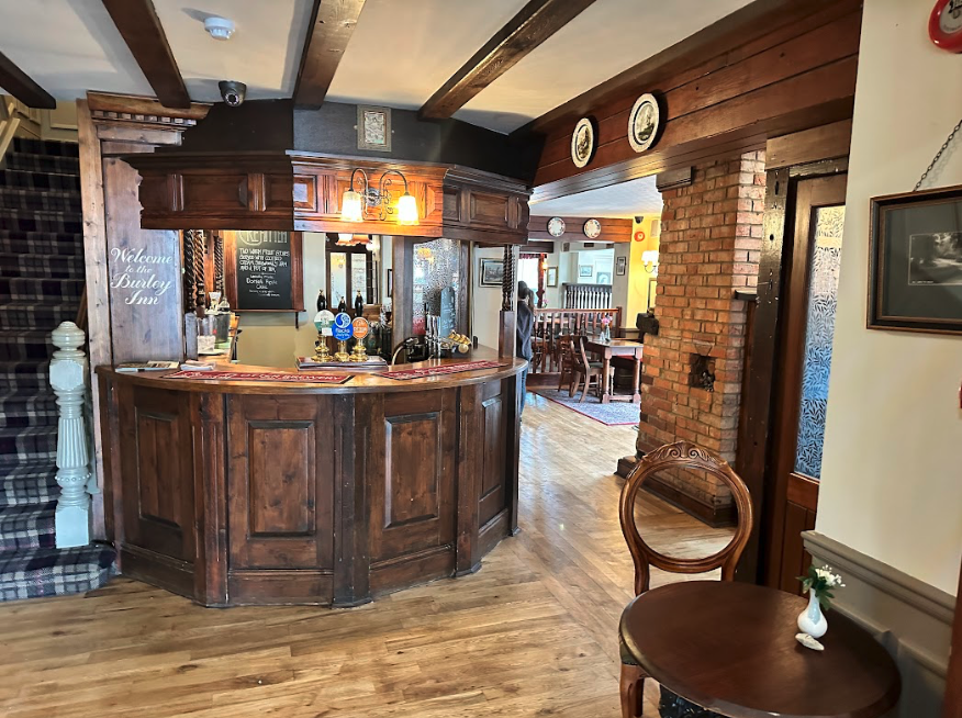 A dark brown pub bar with a sign reading "Welcome to the Burley Inn" next to a plaid staircase.