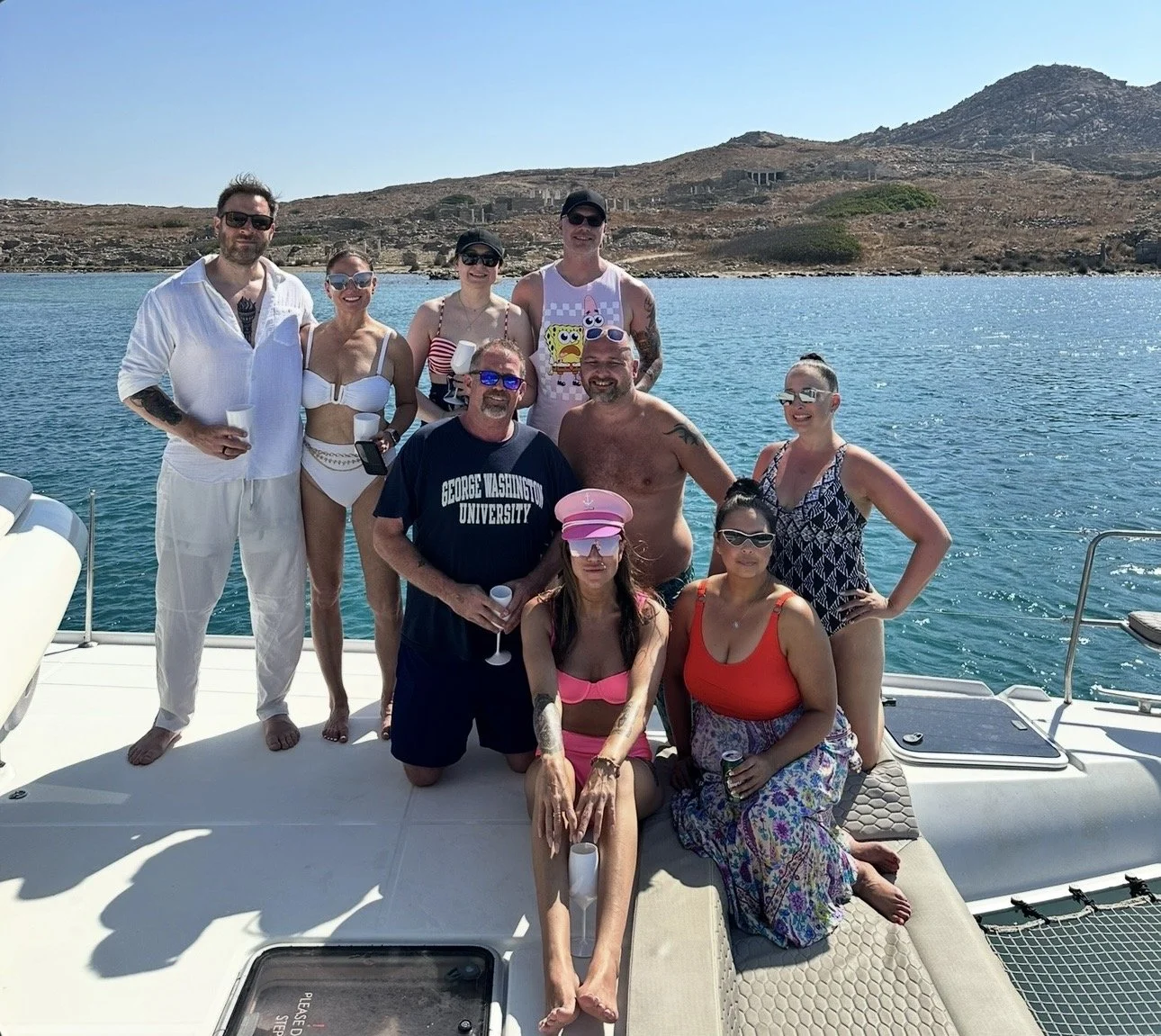 A group of people pose together on a boat deck, in front of blue water and a brown landscape.