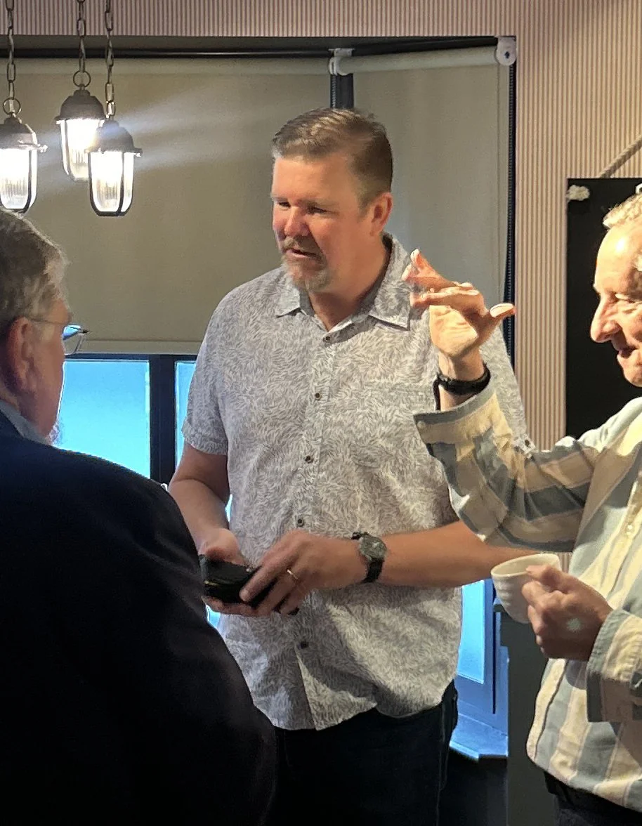 Three men are engaged in conversation indoors; one man in the middle is holding a phone, and the man on the right is holding a bowl and gesturing with his hand. The scene is lit by hanging lamps.