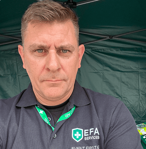 A man wearing a dark polo shirt with EFA Services logo and a green lanyard, standing in front of a green tent at an outdoor event.