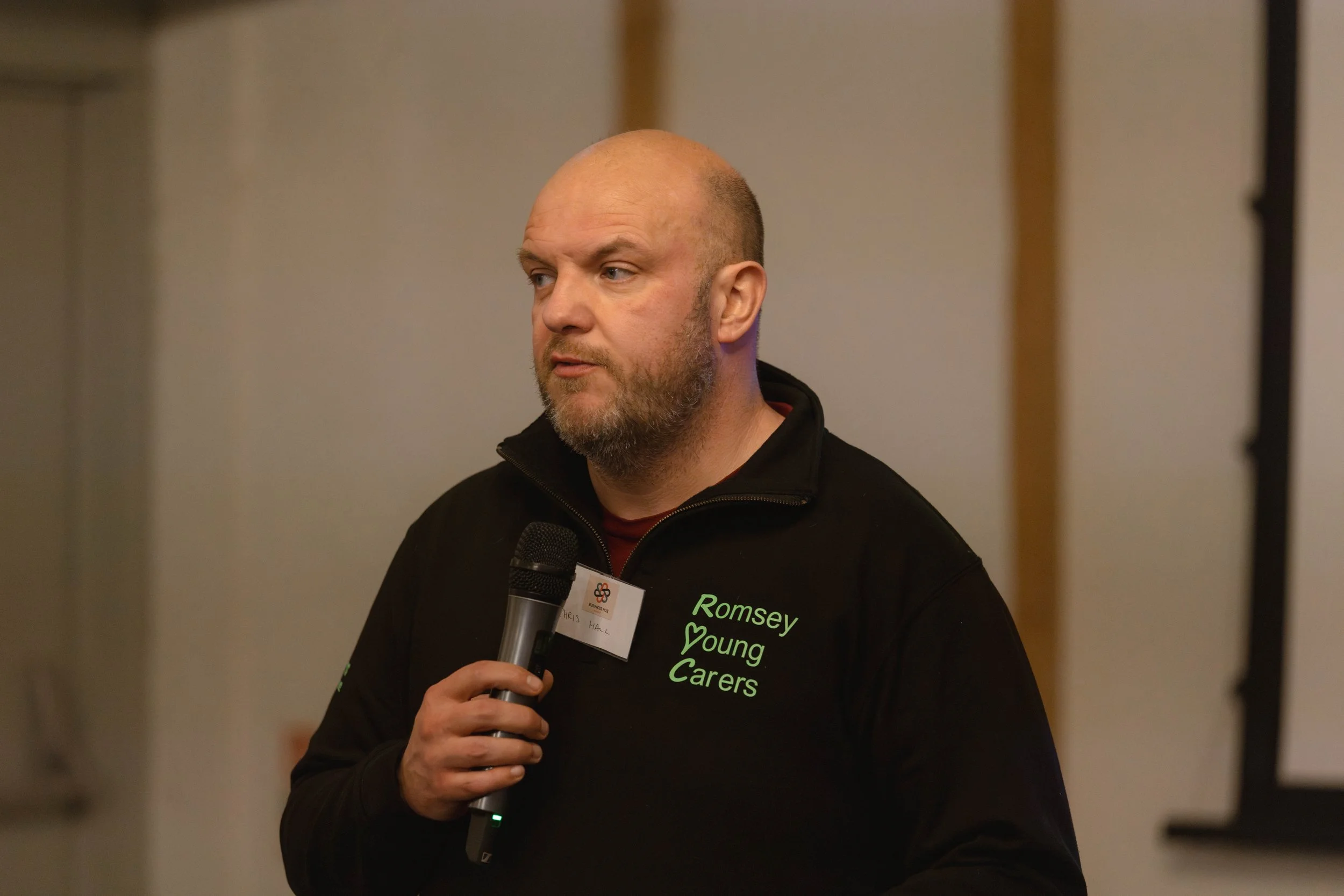 Man with a beard and bald head speaking into a microphone, wearing a black jacket with green text that reads "Romsey Young Carers."