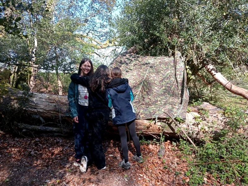 Three young women in outdoor jackets standing in front of a camouflage burlap-covered object or shelter, with leaves and trees in the background.