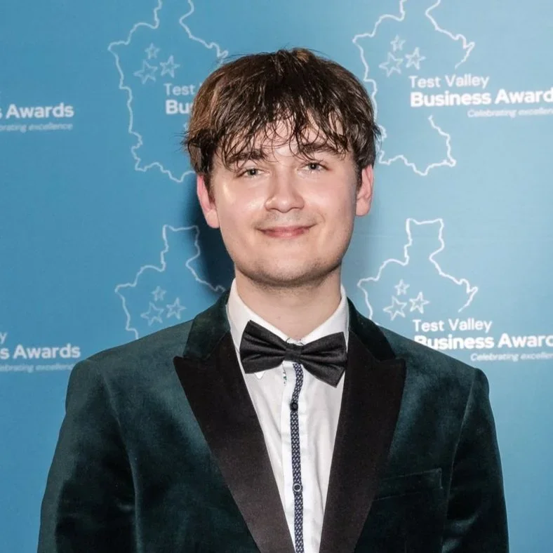Young man in black tuxedo with a bow tie, standing in front of a blue backdrop with text that reads "Test Valley Business Awards" and a map outline, smiling in a formal setting.