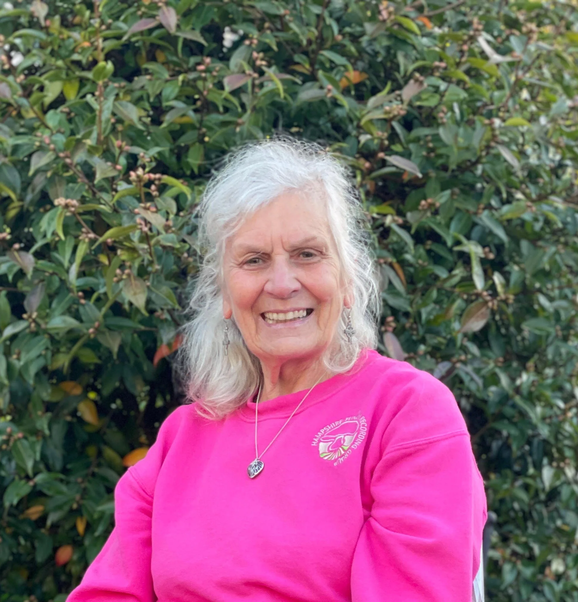 An elderly woman with gray hair, smiling, wearing a pink sweatshirt with a logo, standing outdoors in front of green leafy bushes.
