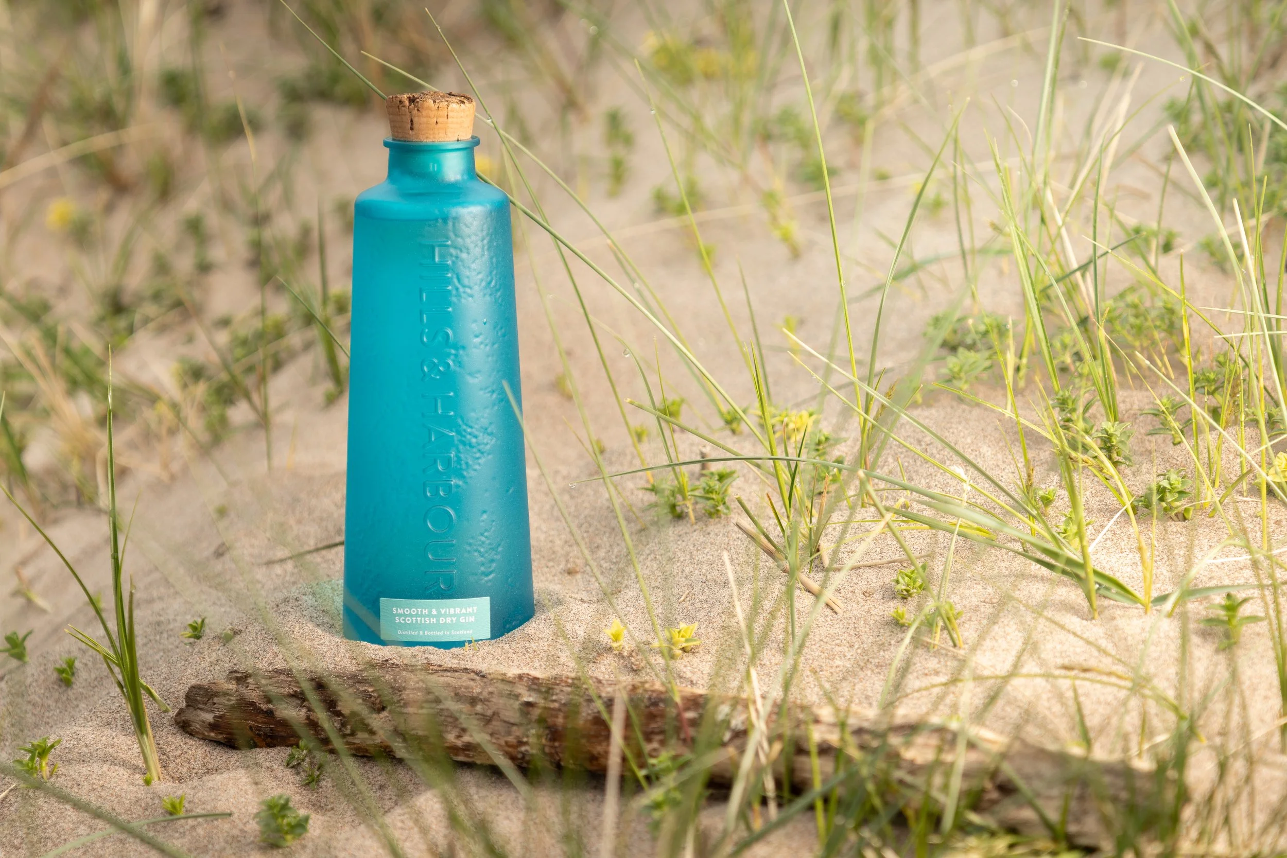 A bottle of Hills and Harbour Gin in the sand with driftwood in front of it and grasses all around it