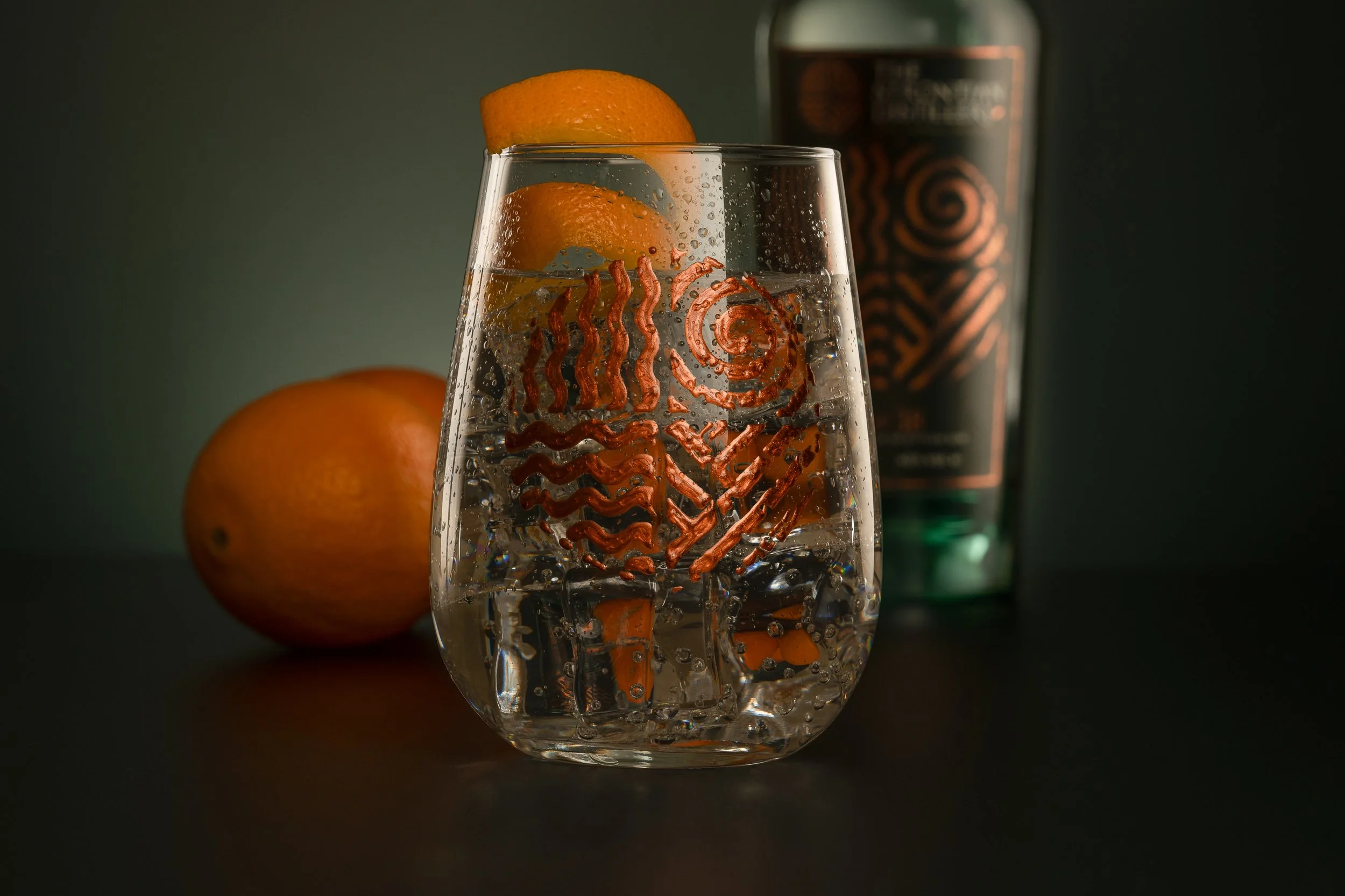Strontian Gin Glass Photograph with orange twist and ice and a Strontian Gin Bottle and oranges in the distance