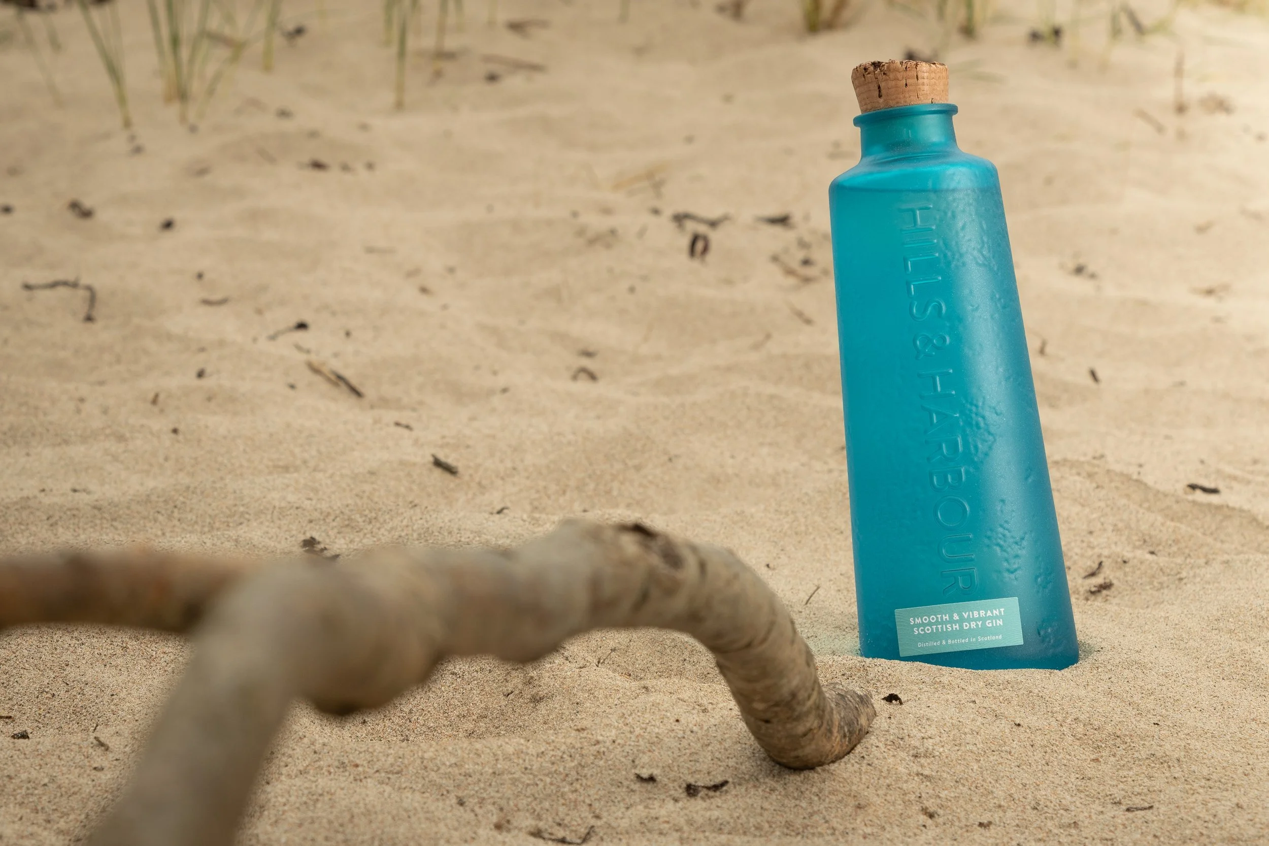 A bottle of Hills and Harbour Gin in the sand with a piece of driftwood in the foreground