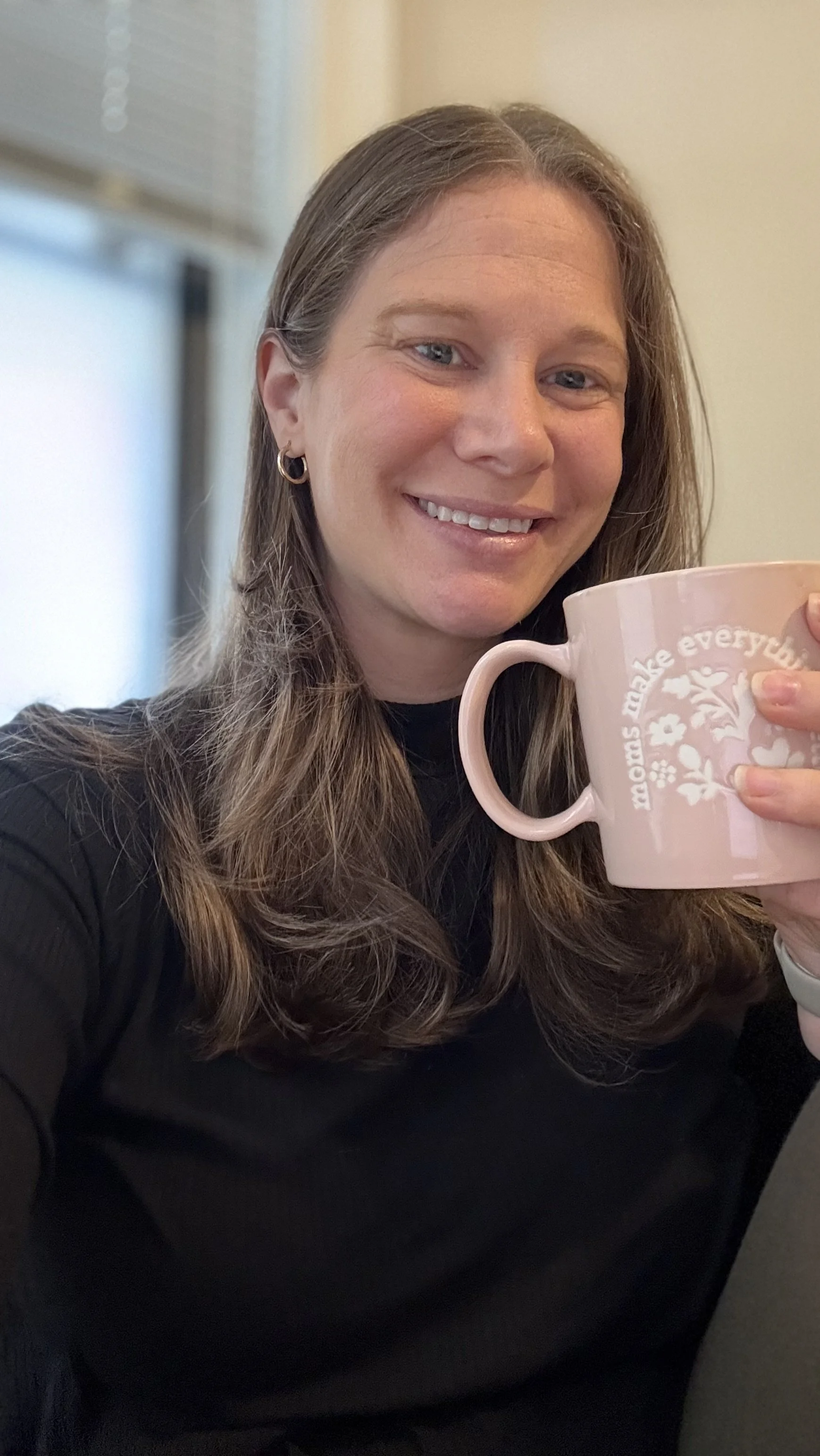 A woman with long brown hair, wearing hoop earrings and a black shirt, smiling and holding a pink mug with a floral design and the text 'moms make everything better' on it.