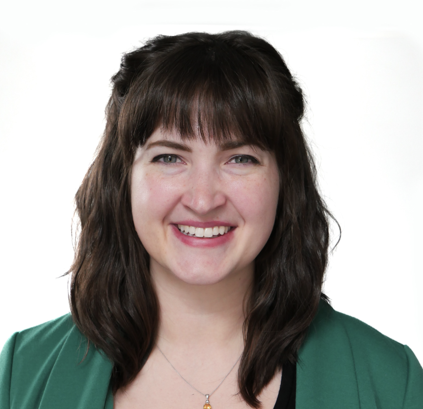 A smiling client with dark brown hair, green eyes, and fair skin, wearing a green blazer and a necklace with a small pendant, against a white background.