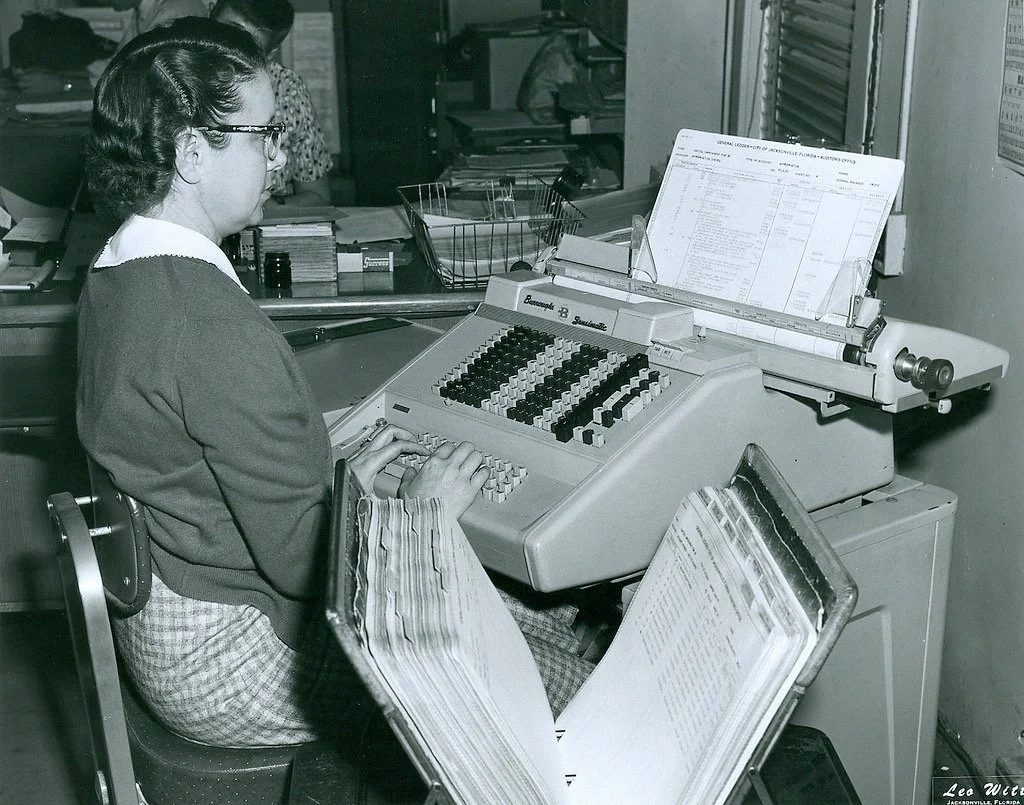 A woman working at a vintage office typewriter with a sheet of paper and a clipboard nearby.