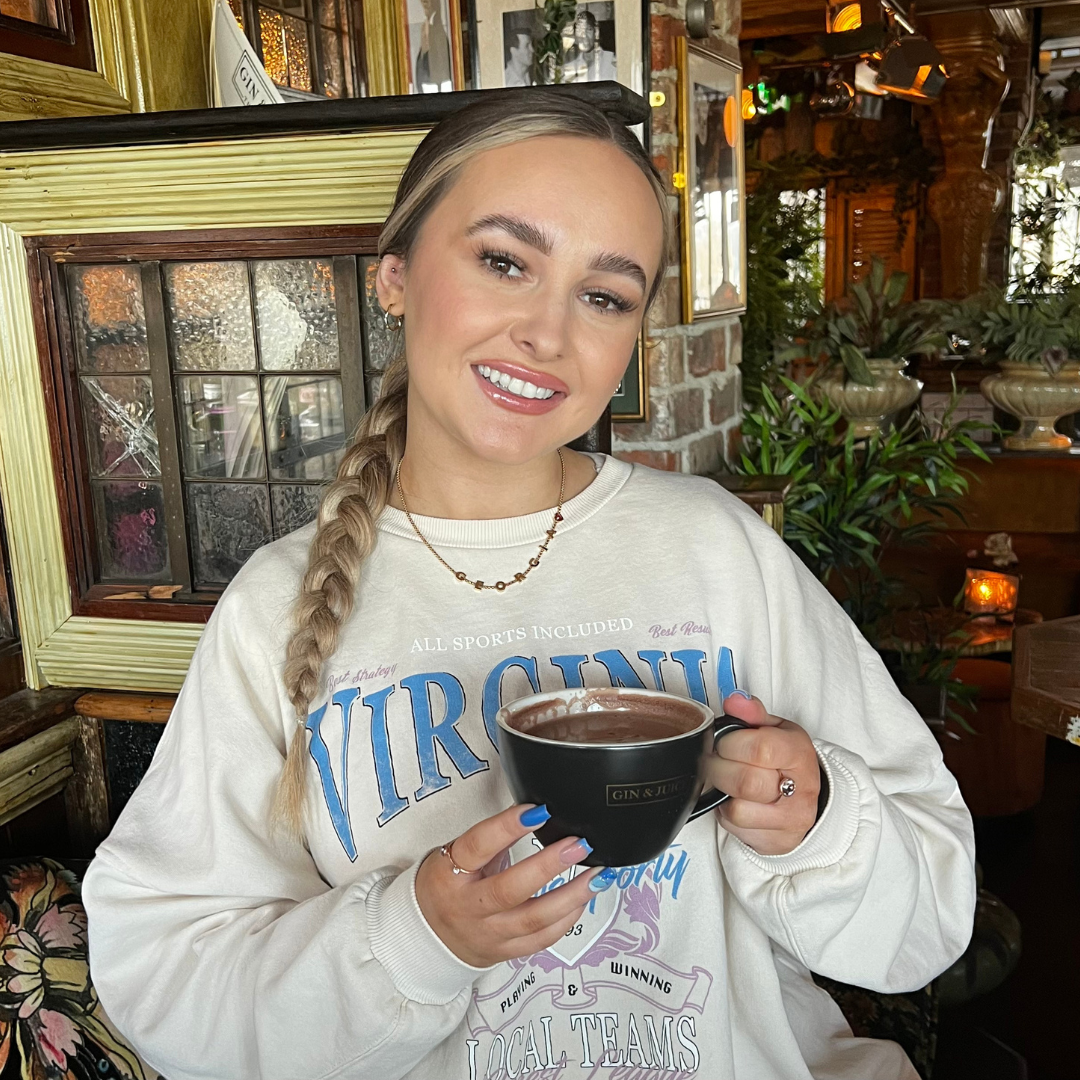 A young woman with long braided hair, light makeup, and a bright smile holds a large black mug filled with a hot beverage in a cozy, decorated restaurant. She wears a white sweatshirt with colorful text and small jewelry, and is sitting in a warmly-lit, rustic interior with plants and framed artwork.
