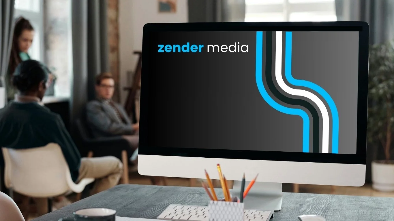 Computer monitor displaying 'zender media' logo in an office setting with three people sitting and working in the background.