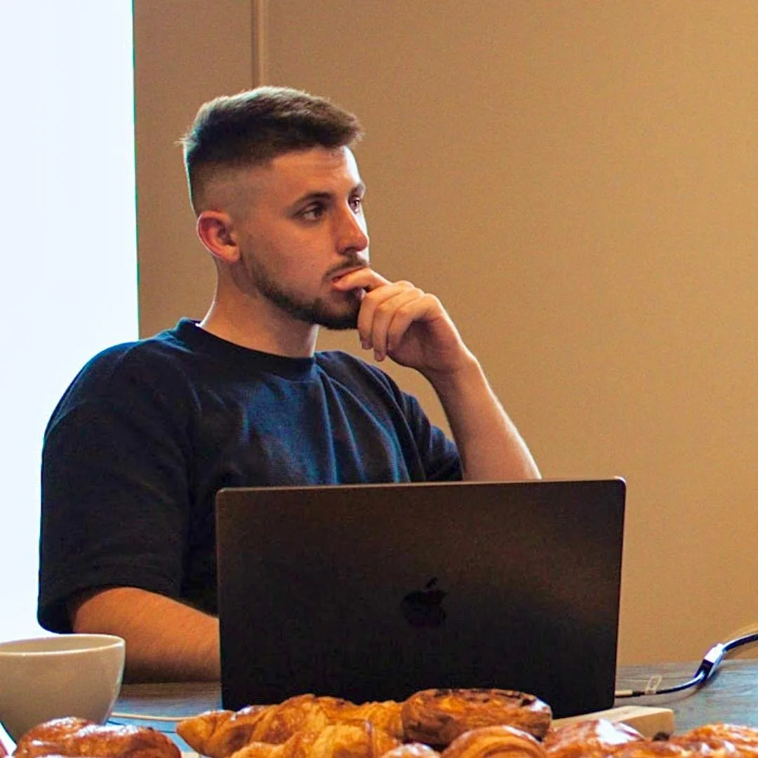 A young man with short hair and a beard, wearing a black T-shirt, sitting at a table with a laptop, eating pastries, and drinking from a white mug.