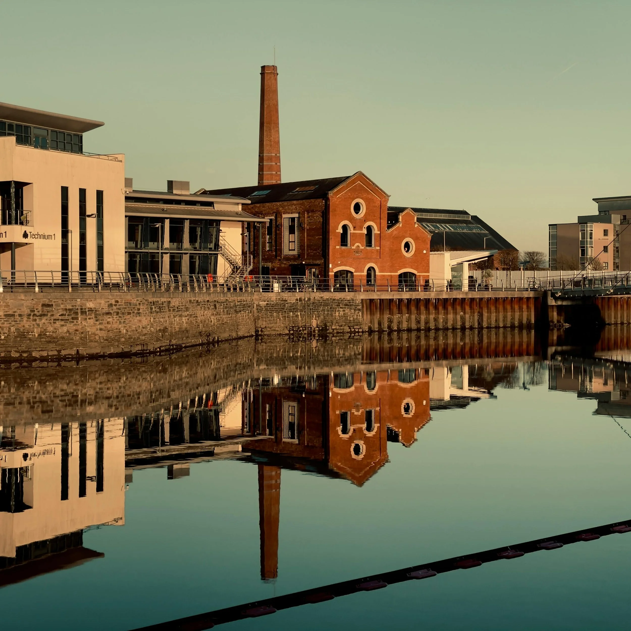 A view of a brick building and industrial factory reflected in a calm body of water during sunset.