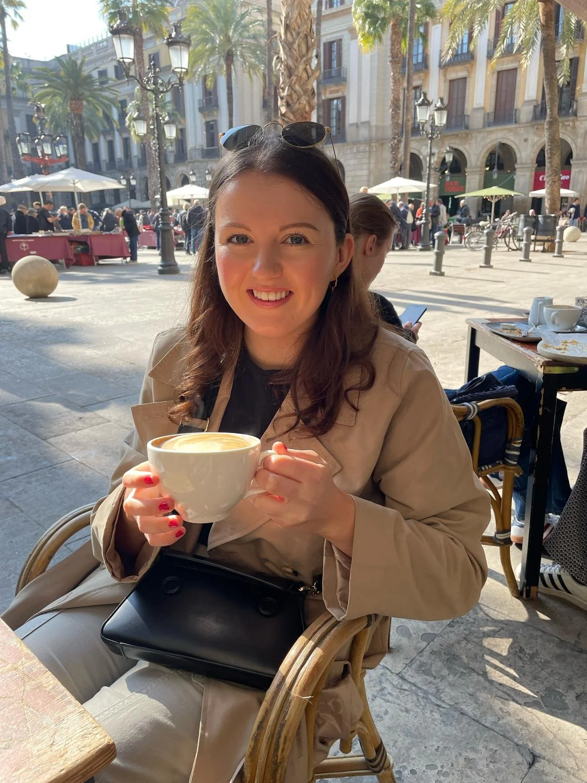 A woman sitting outdoors at a café holding a large cup of coffee, smiling at the camera. In the background, there are palm trees, umbrellas, and people walking in a city square with a building that has balconies.