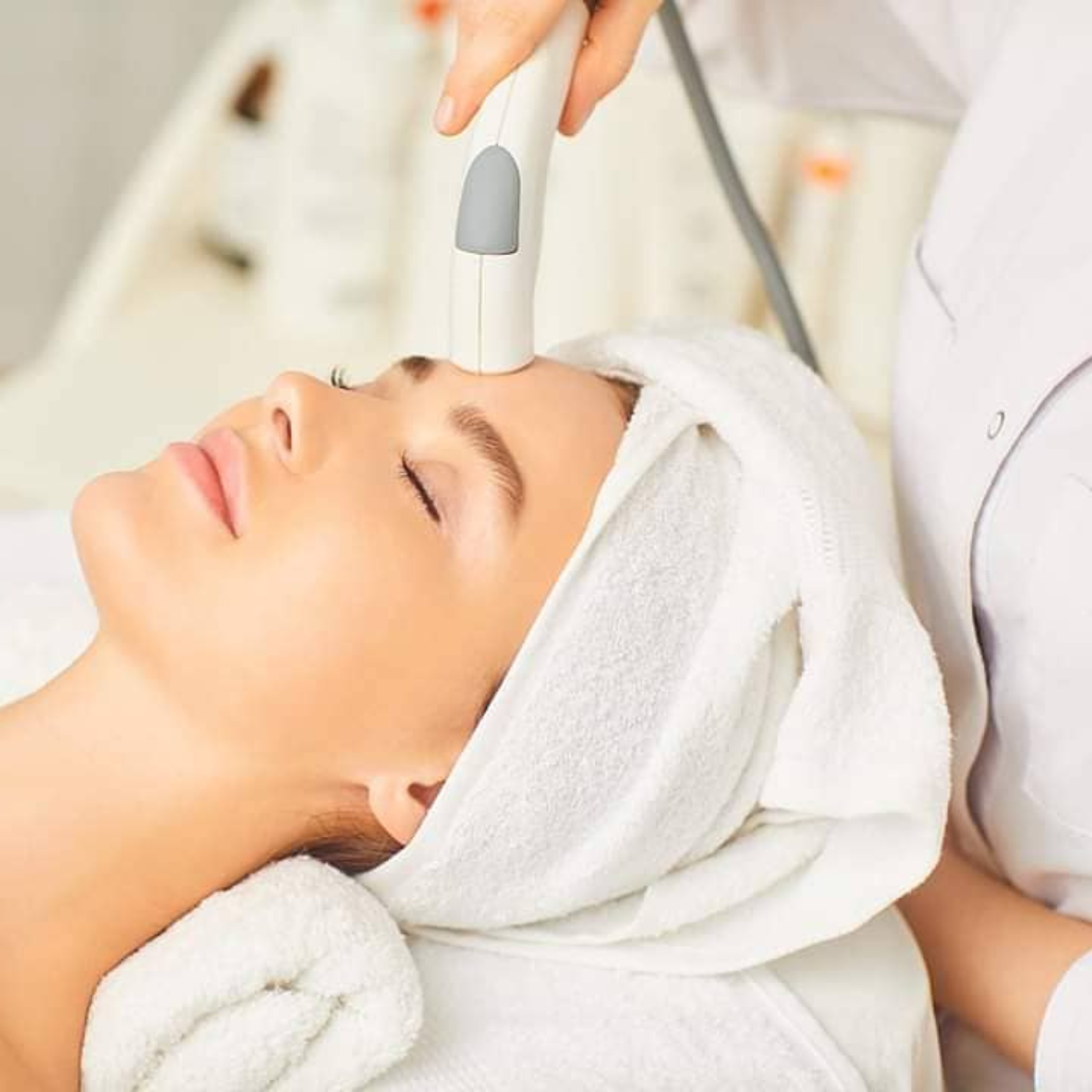 A woman receiving a diamond e-360 facial treatment with a handheld device at a spa or skincare clinic, lying on a treatment bed with a towel wrapped around her head.
