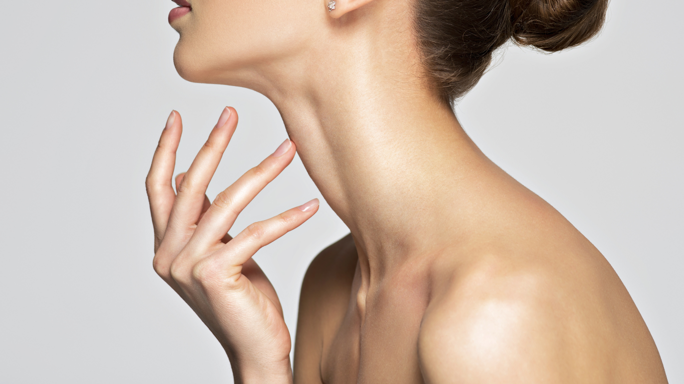 Close-up of a woman's neck, chin, and shoulder with smooth skin, touching her neck with her fingers, against a neutral background.