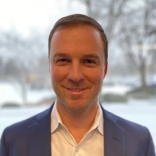 A man with short brown hair and a beard, smiling, wearing a dark blue suit jacket and white shirt in front of a window with a snowy landscape outside.