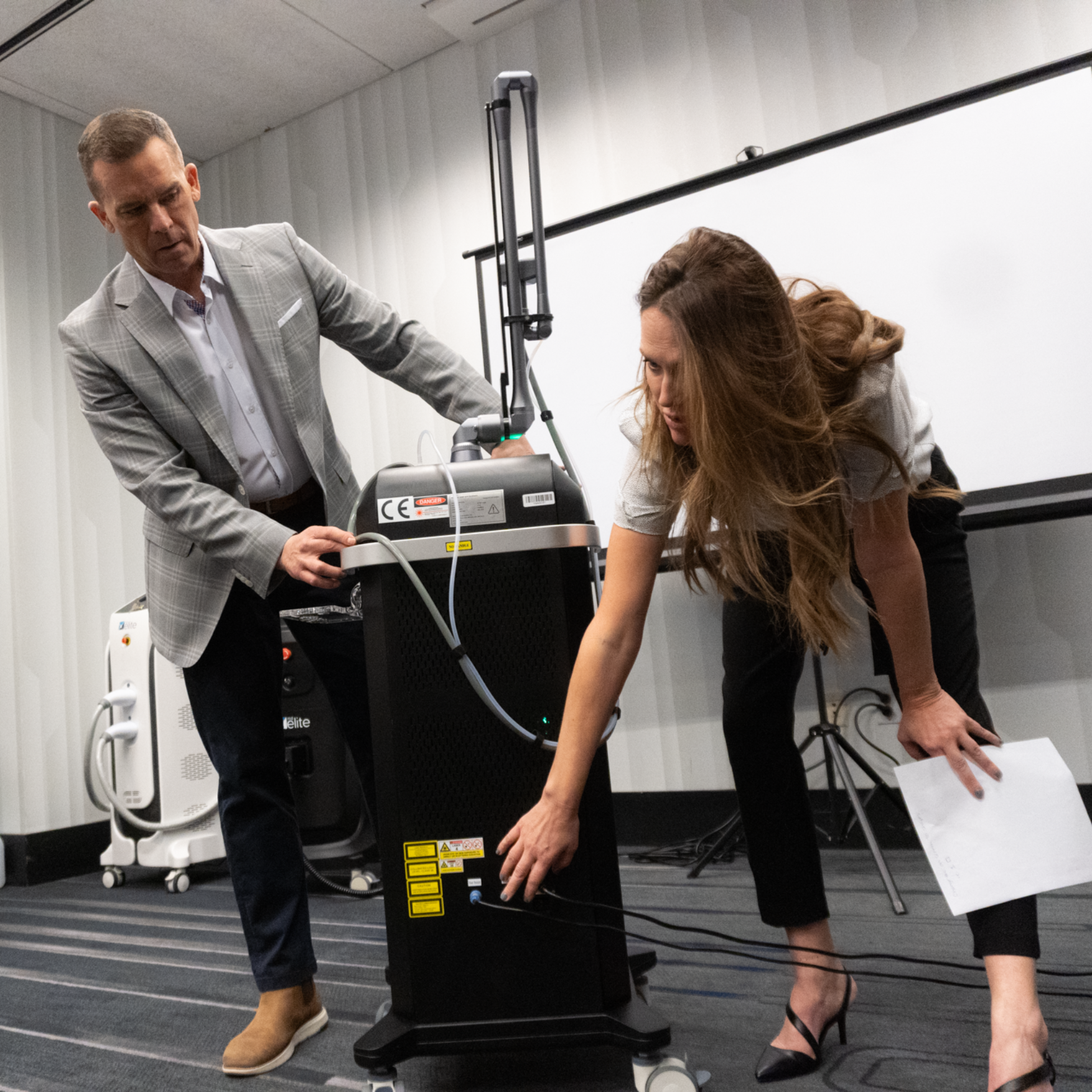 A man in a plaid blazer and a woman in a white top and black pants are operating medical or scientific equipment in a room with a whiteboard. The woman is adjusting a cable or connection on a black tower-like device, while the man observes.