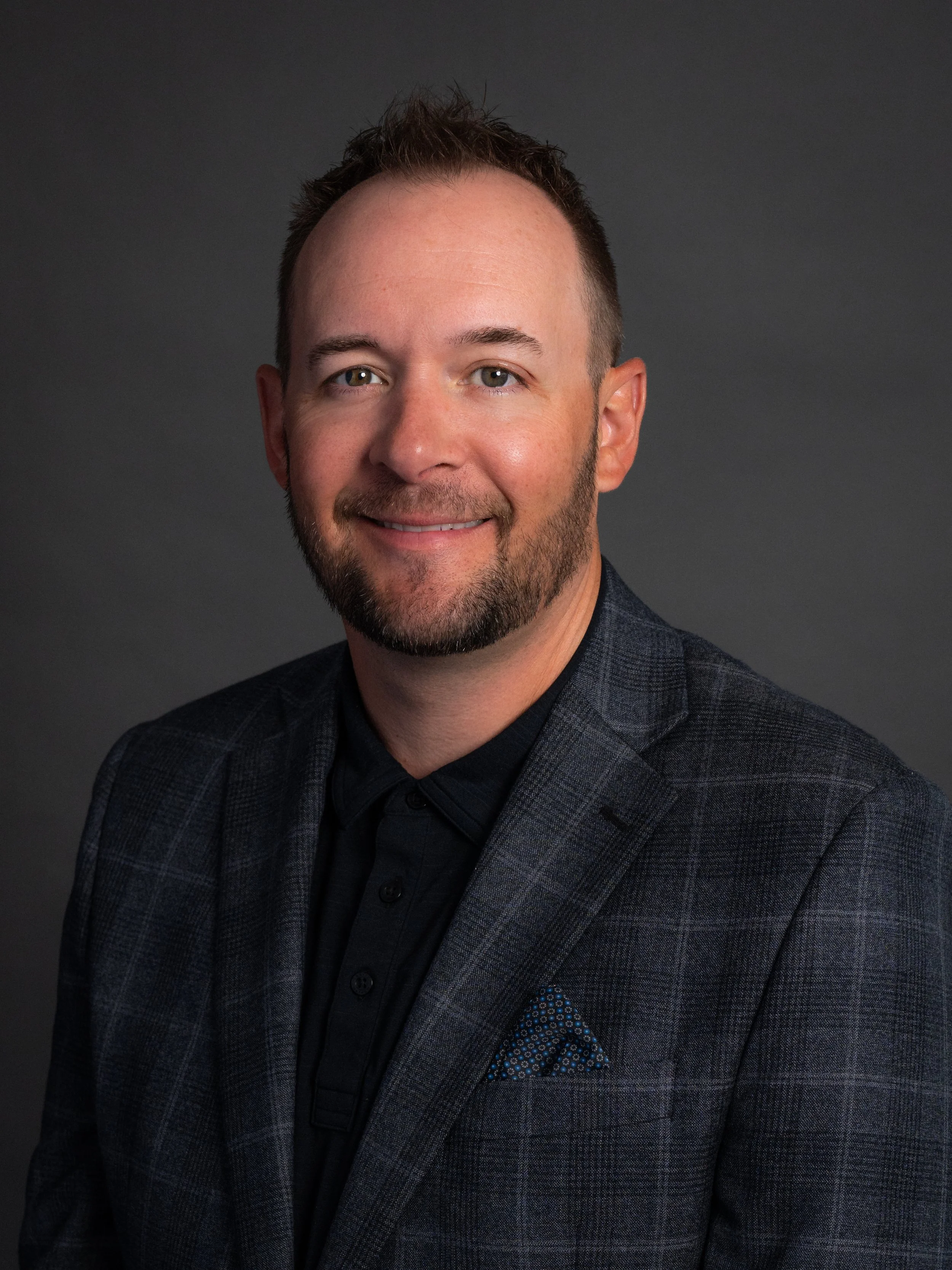 Headshot of Blair with light skin, short brown hair, a beard and mustache, wearing a dark checked blazer over a black shirt, smiling against a dark gray background.