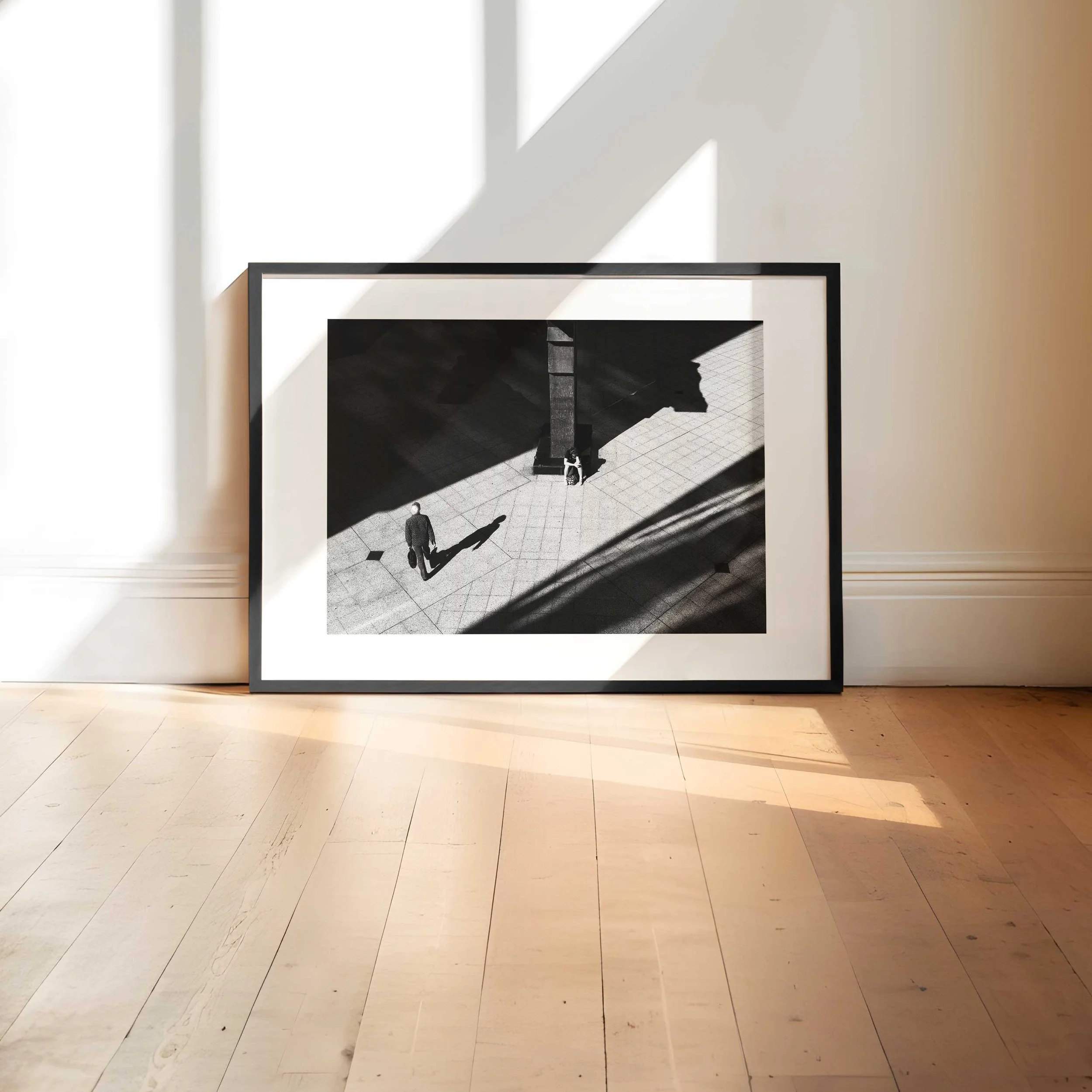 Black and white street photograph of two isolated figures separated by strong geometric shadows and light inside a modern architectural space.