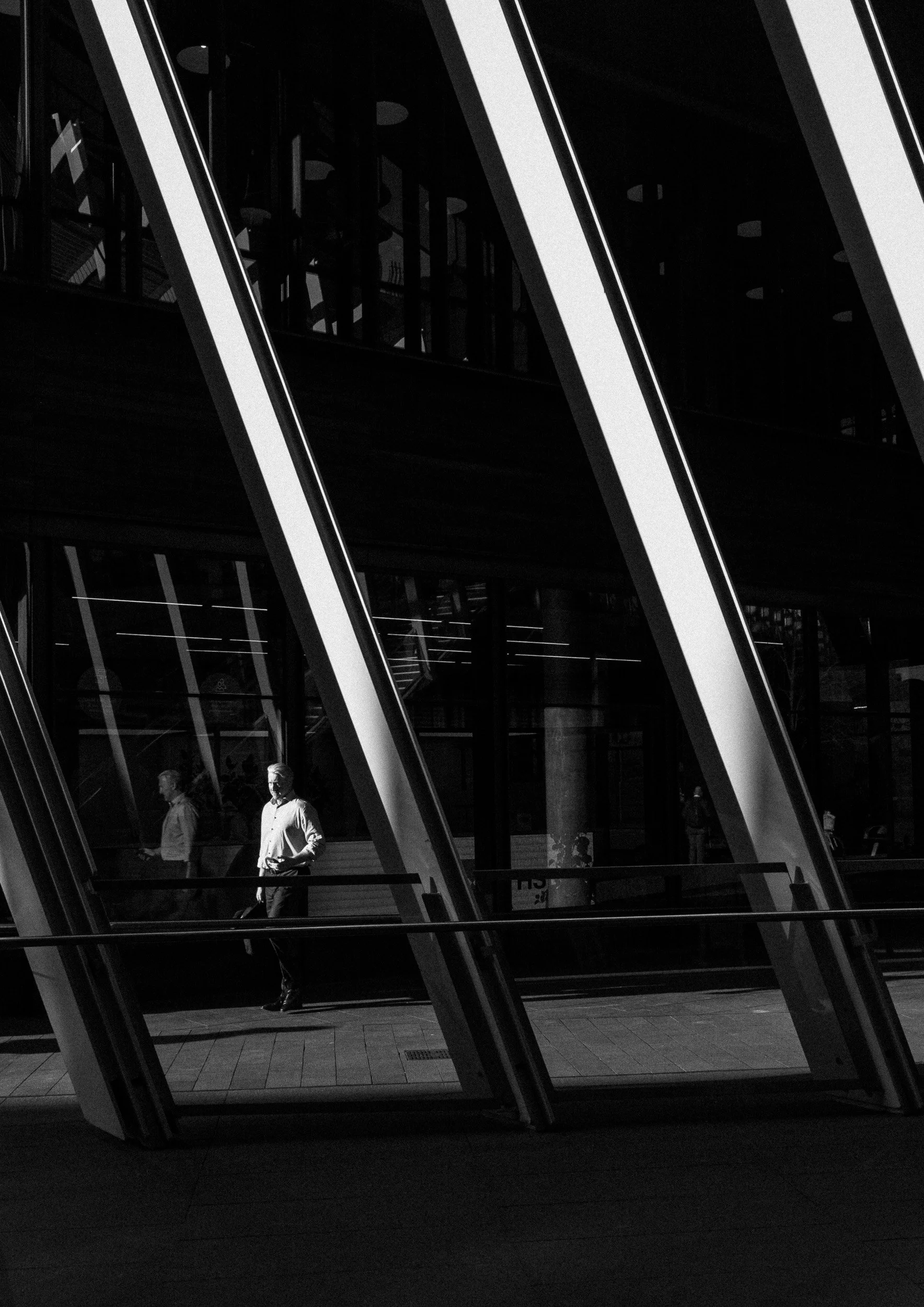 Black and white street photograph of a man walking across diagonal architectural lines with reflections and geometric composition