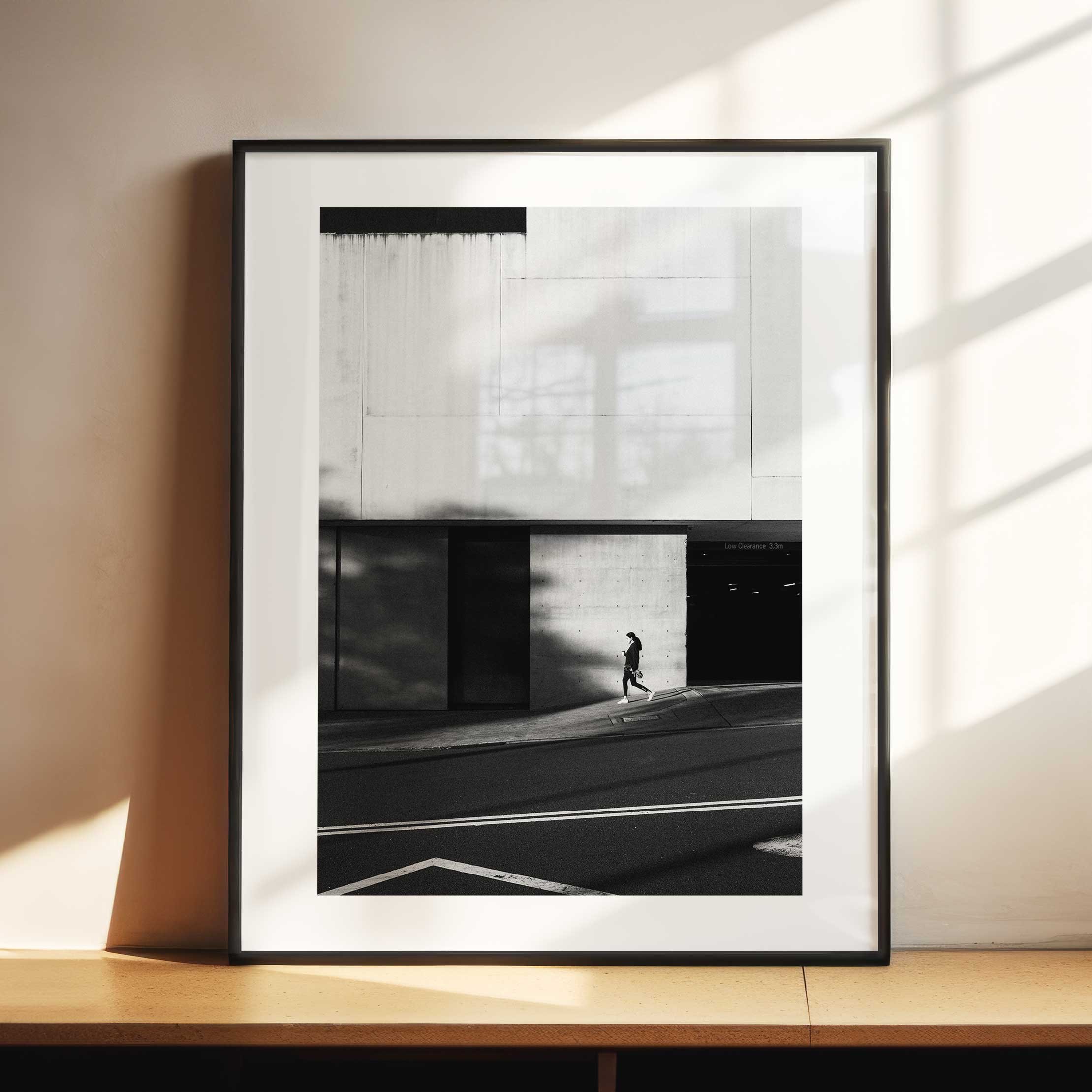 Black and white street photograph of a lone pedestrian walking through a shaft of light beneath a large geometric concrete facade in Sydney.