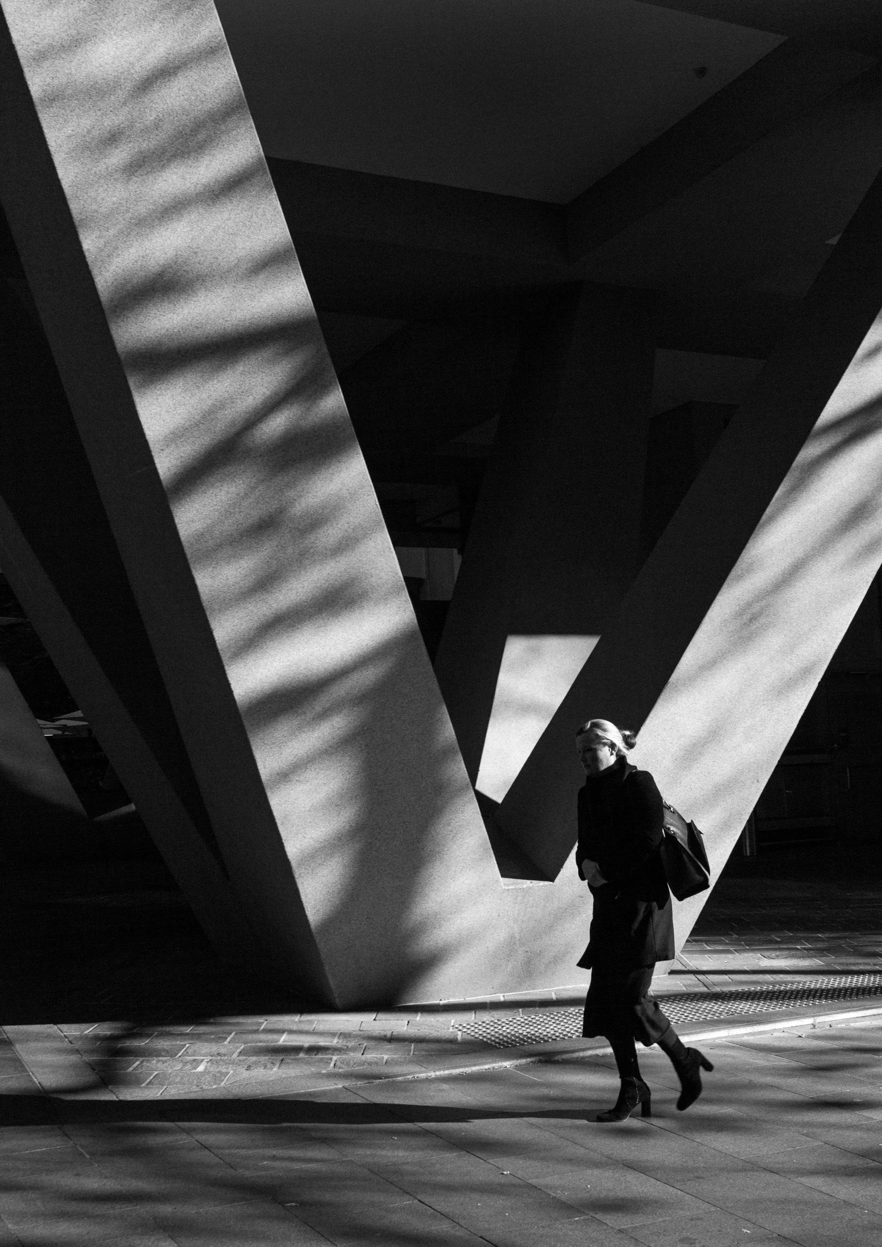 Black and white street photograph of a person walking through strong geometric architectural structures with light and shadow contrast