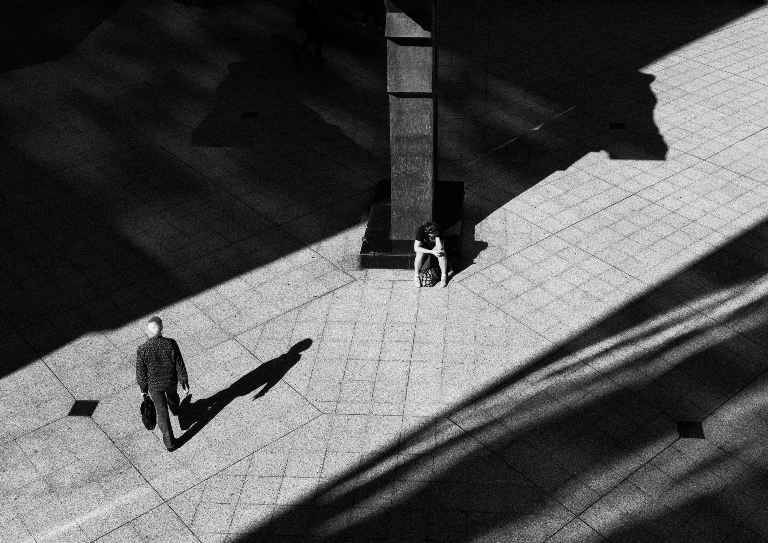 Black and white street photograph of two isolated figures separated by strong geometric shadows and light inside a modern architectural space.