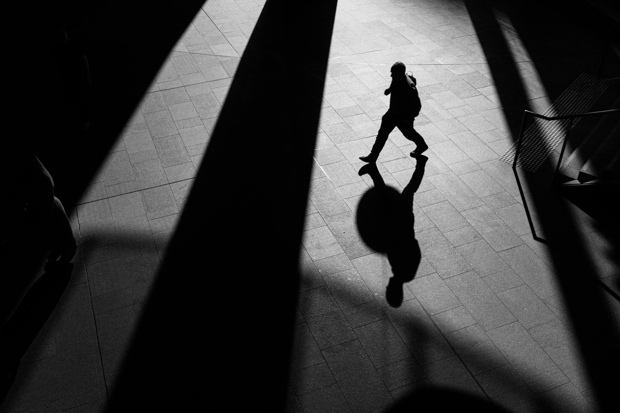Black and white street photography of a person walking with a long shadow across a bright surface with strong light contrast