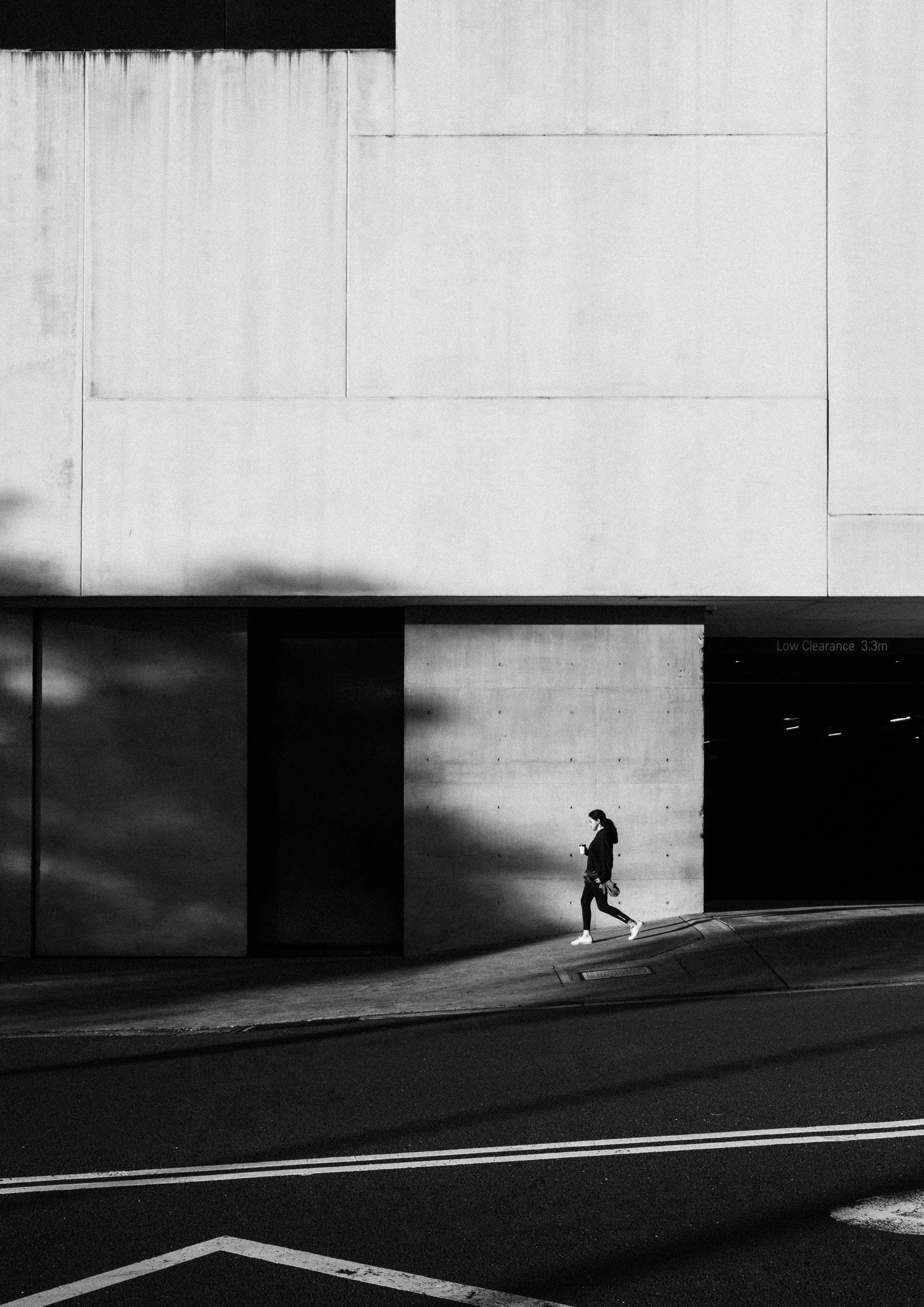 Black and white street photograph of a lone pedestrian walking through a shaft of light beneath a large geometric concrete facade in Sydney.