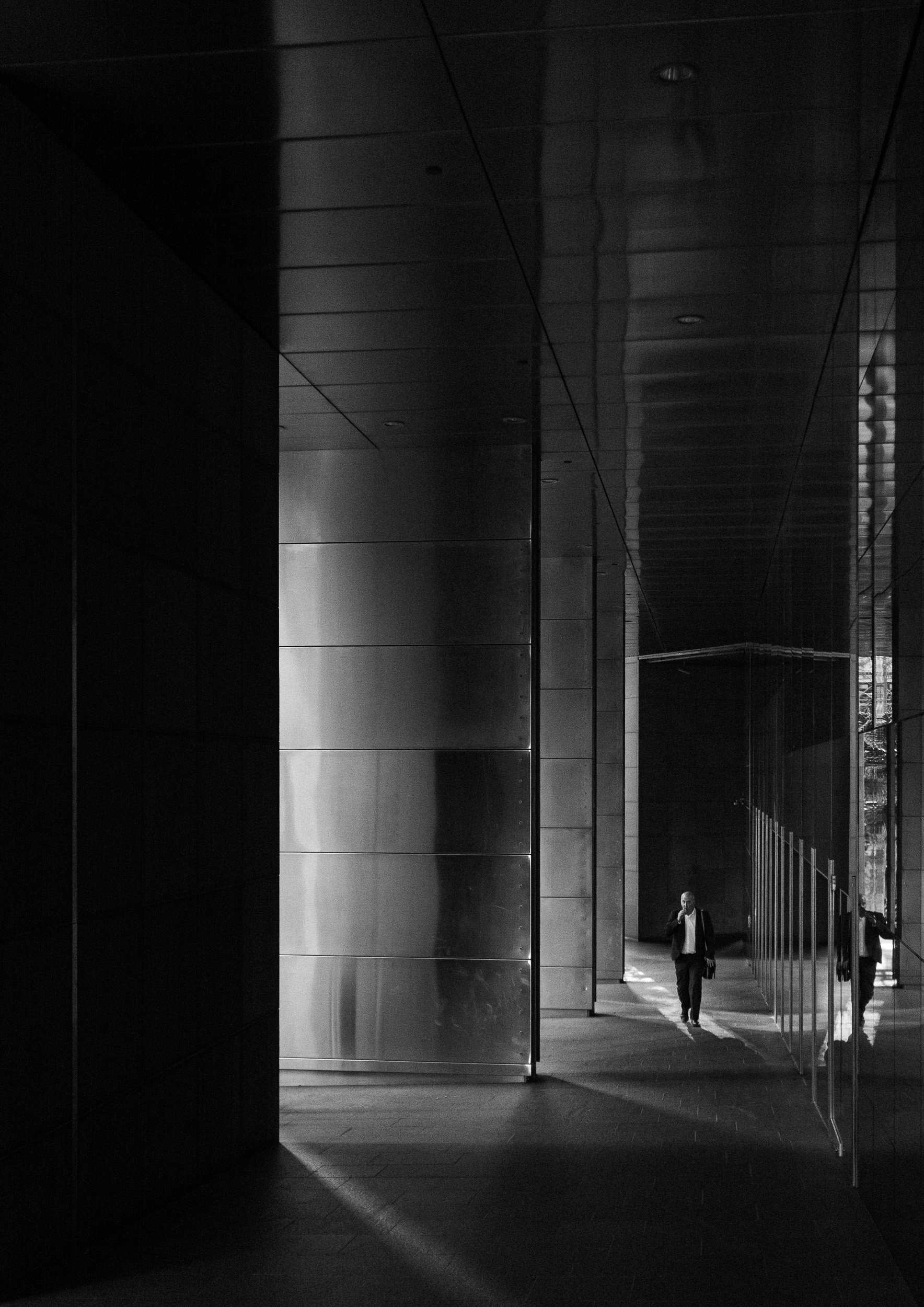 Black and white architectural street photography of a man walking through a narrow corridor with reflections and strong vertical lines