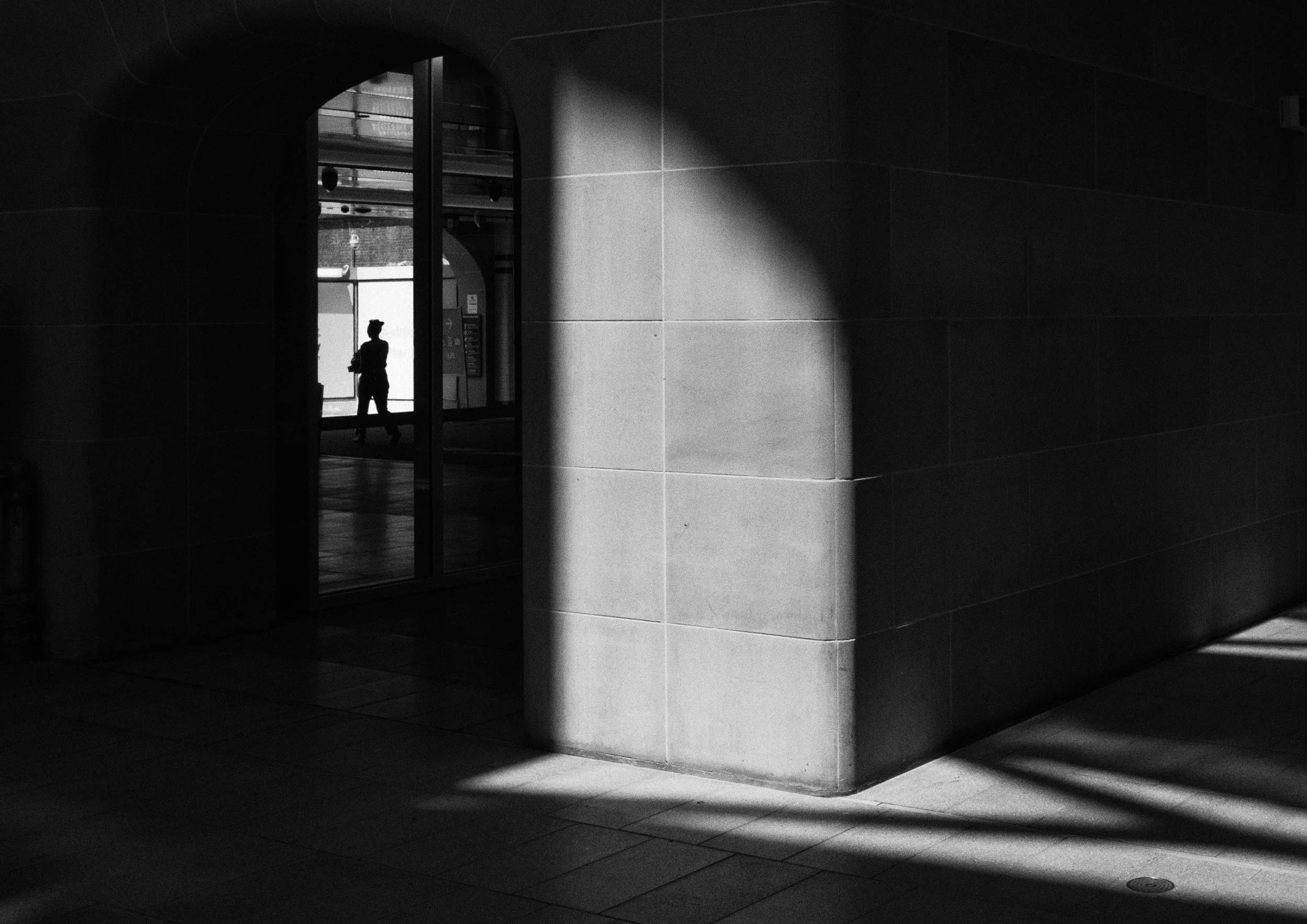 Black and white street photograph of a person walking through a glass doorway framed by strong light and shadow, with geometric architectural lines and high contrast lighting.